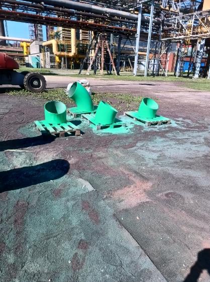 Cztery zielone rury na paletach po malowaniu, na tle instalacji przemysłowej. Widoczne ślady farby na podłożu, co sugeruje niedawne malowanie.