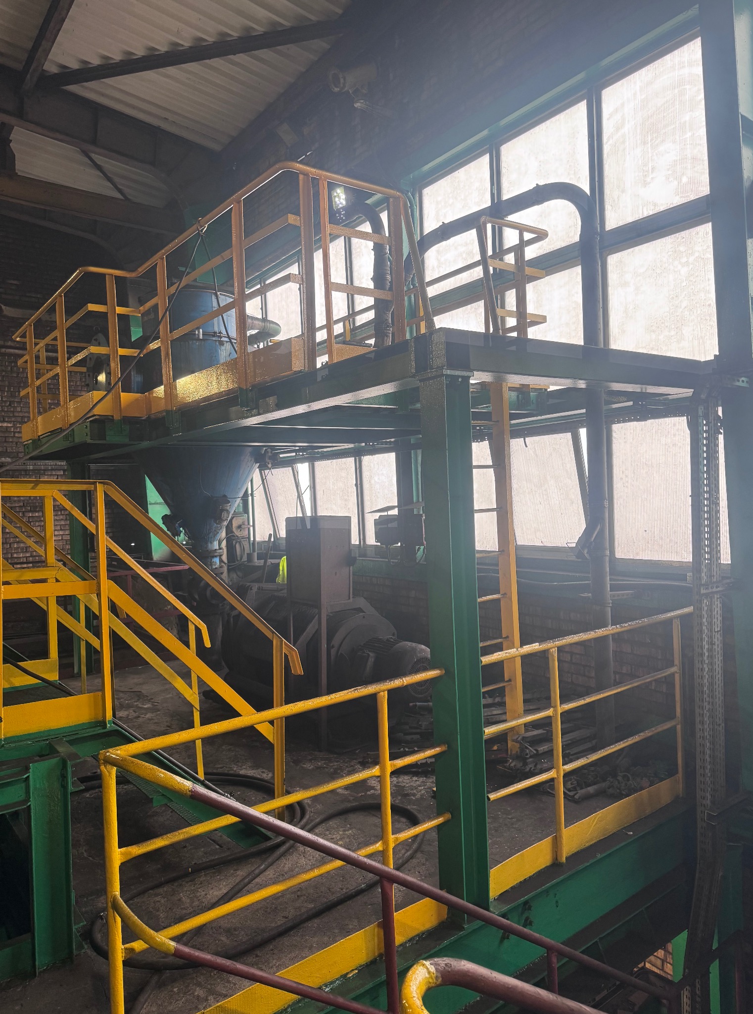 Wnętrze hali przemysłowej z metalową konstrukcją pomalowaną na zielono i żółto, widoczny fragment instalacji z zbiornikiem, okna w tle.