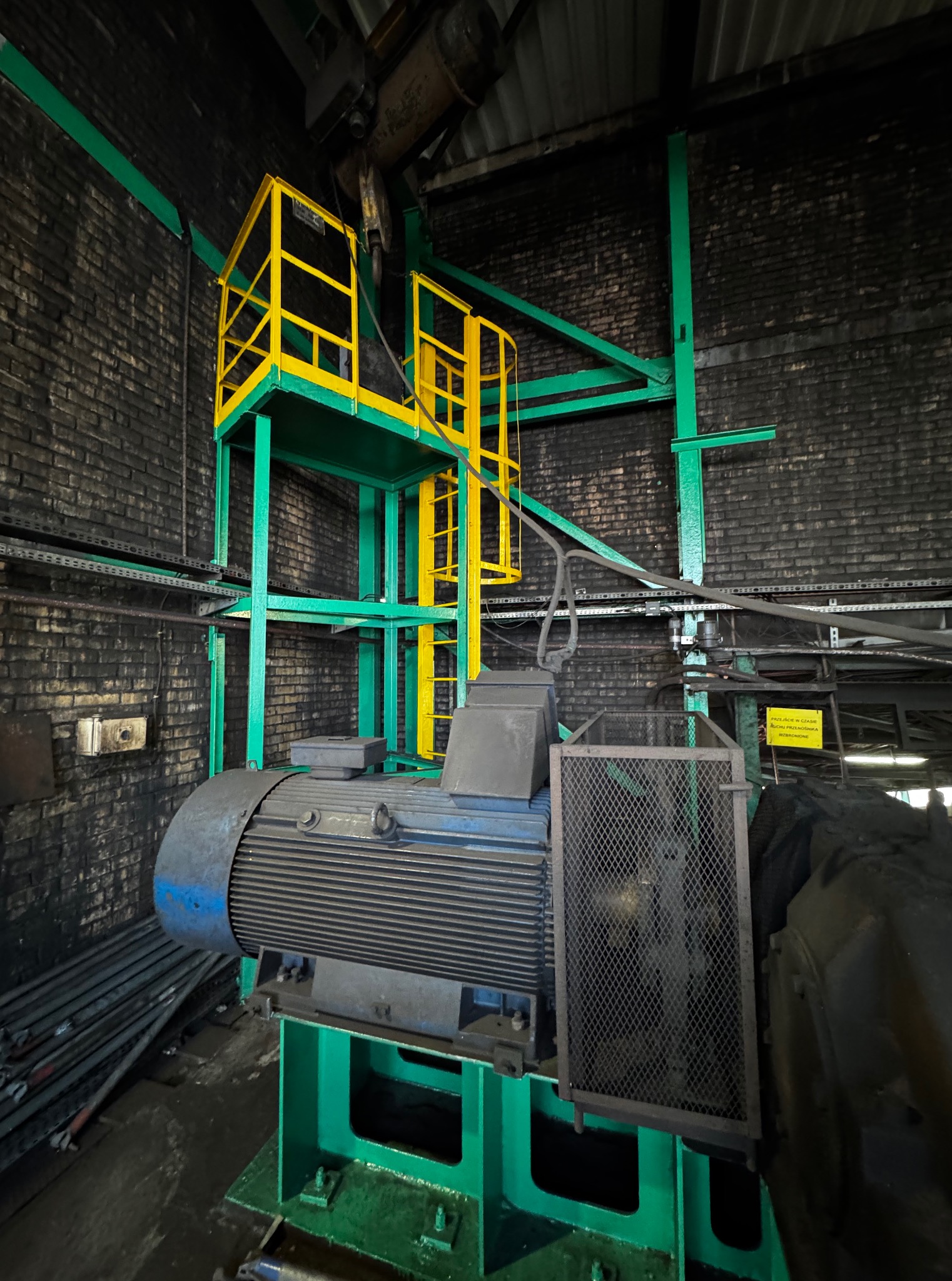 Wnętrze hali przemysłowej z ciemną, zabrudzoną cegłą i fragmentem maszyny z metalową kratą. Konstrukcja pomalowana na zielono i żółto. Widoczne ślady eksploatacji.