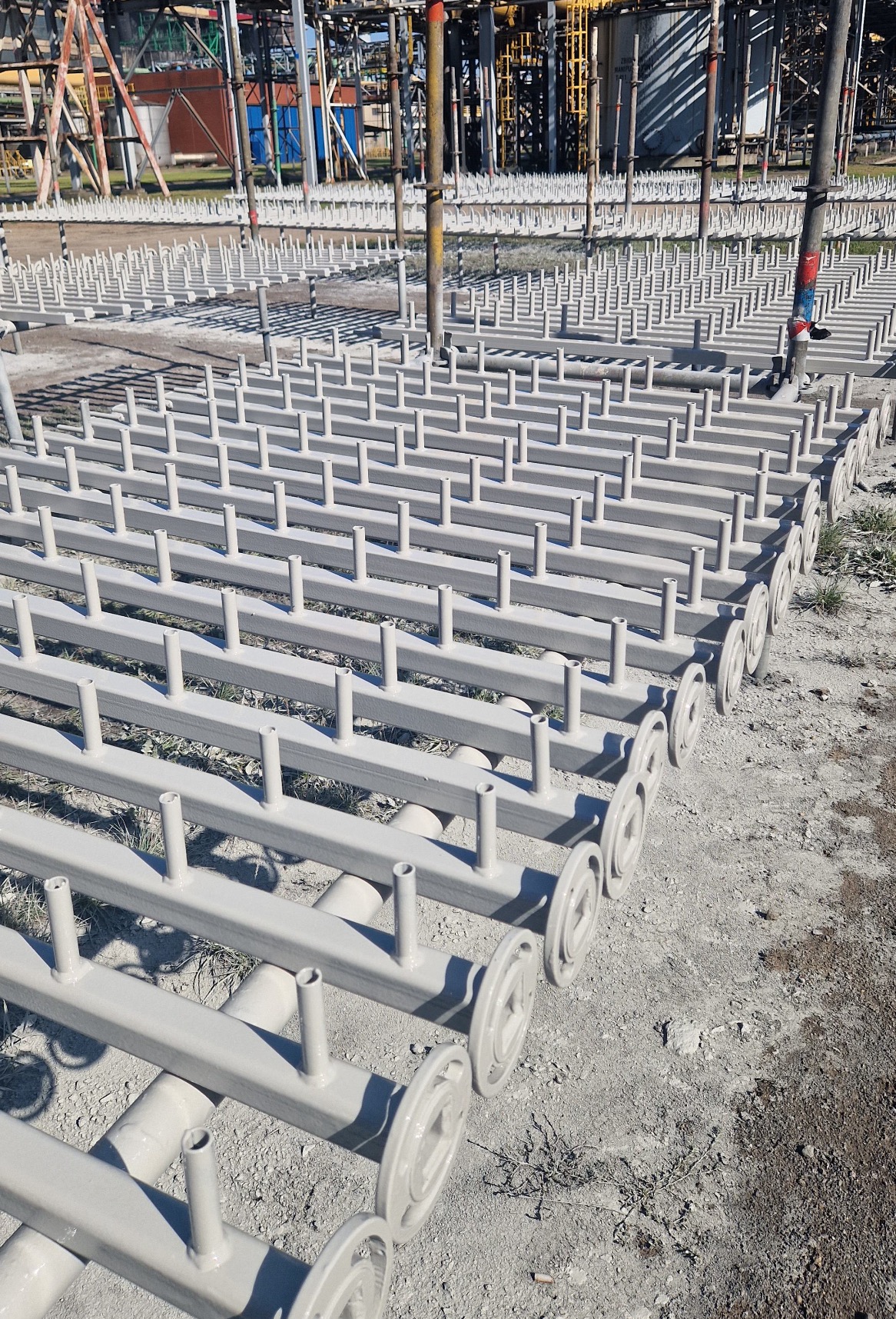 Metalowe konstrukcje po obróbce, pokryte jednolitą szarą farbą, ułożone rzędami na zewnątrz, z widoczną infrastrukturą przemysłową w tle.