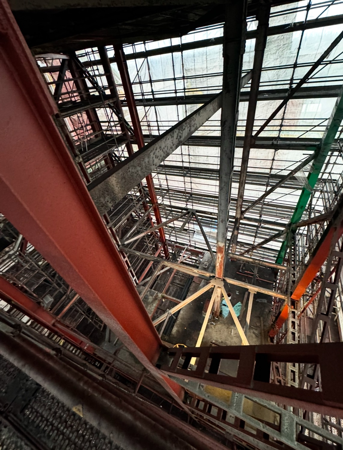 Wysoka konstrukcja stalowa z rusztowaniami, widok z dołu. Elementy pomalowane na czerwono, szaro i zielono. Widoczne zacieki i ślady korozji.