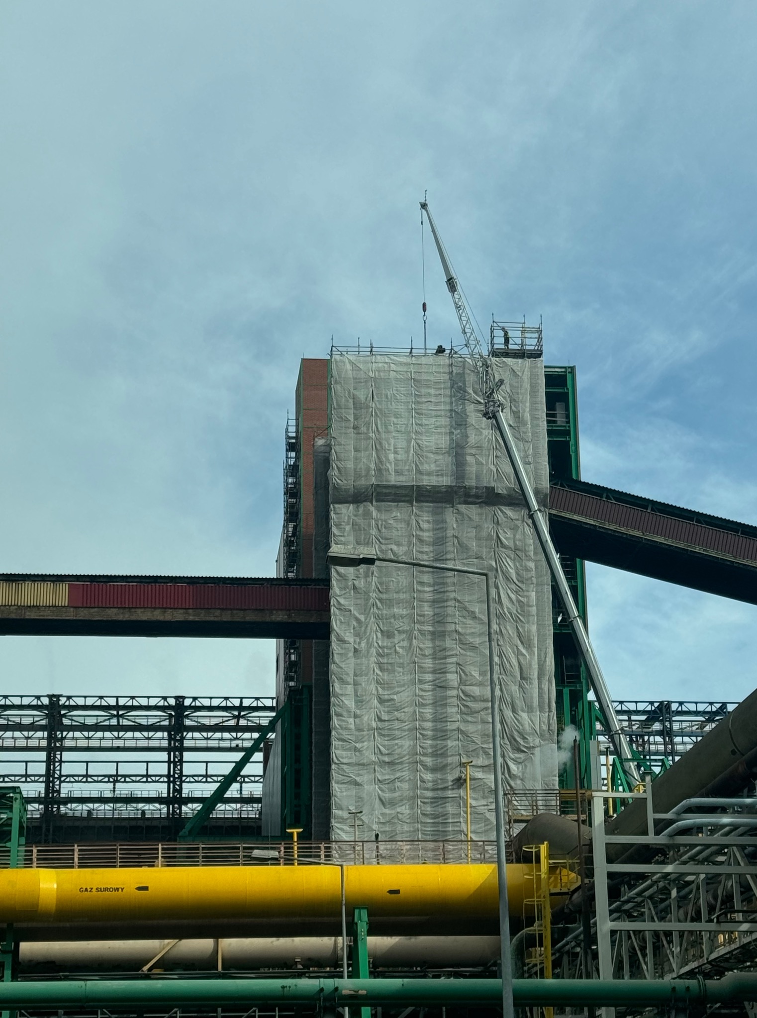 Przemysłowy budynek w trakcie prac renowacyjnych, osłonięty siatką, z dźwigiem i widocznymi rurociągami. Elementy konstrukcyjne w kolorach zielonym i żółtym.