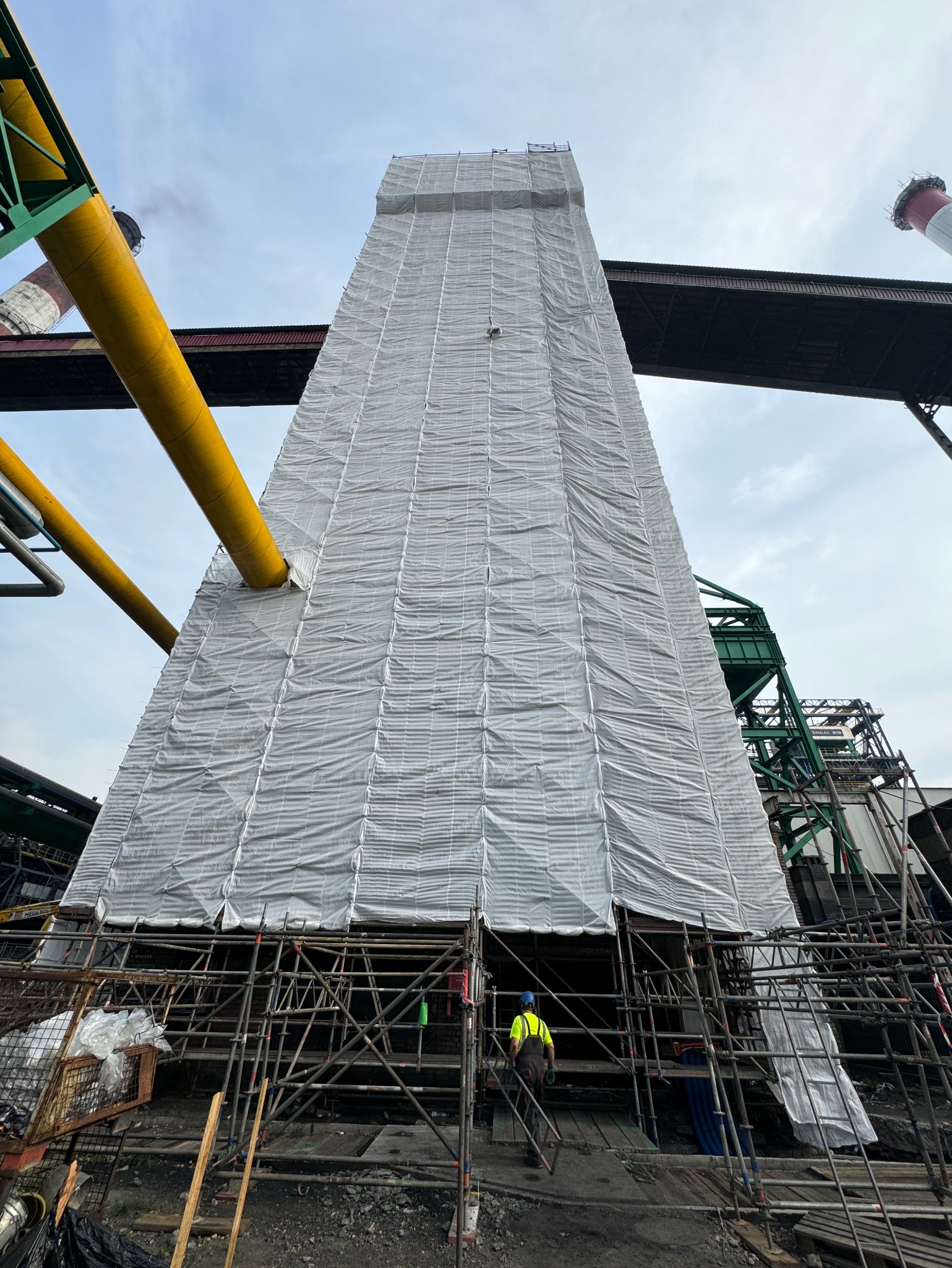 Wysoka konstrukcja przemysłowa osłonięta siatką ochronną na rusztowaniu, widoczny pracownik w stroju roboczym, fragmenty instalacji i kominów w tle.