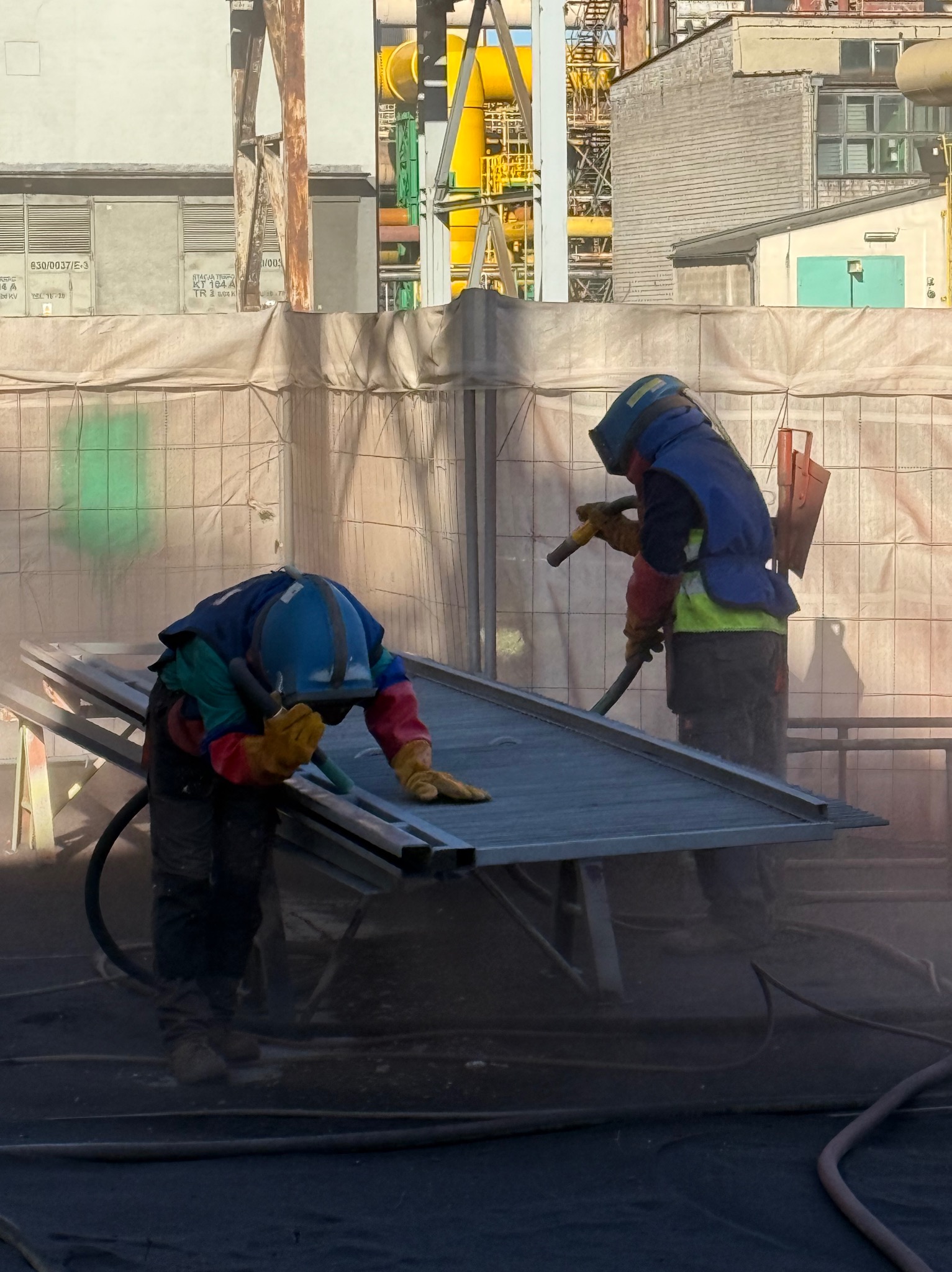 Dwóch pracowników w kombinezonach ochronnych czyści metalową konstrukcję za pomocą specjalistycznego sprzętu na tle przemysłowego krajobrazu.