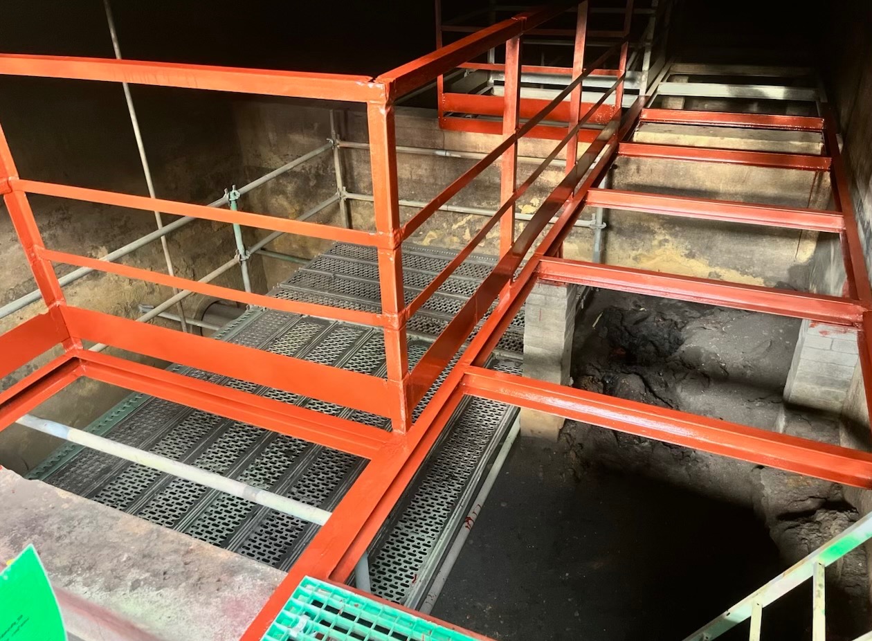 Metalowa konstrukcja schodów z pomalowanymi na pomarańczowo barierkami i kratami, prawdopodobnie po malowaniu proszkowym w przestrzeni przemysłowej.