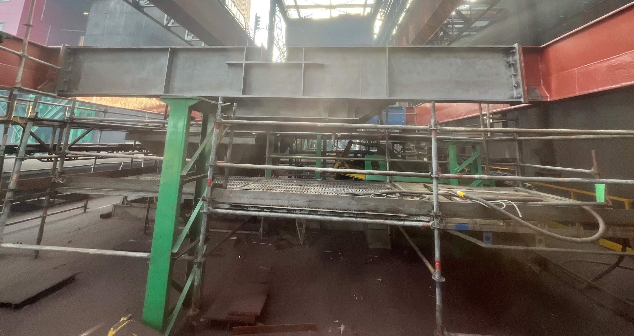 Konstrukcja stalowa na rusztowaniu, częściowo pomalowana na czerwono, w hali przemysłowej. Widoczne spawy i elementy mocujące.