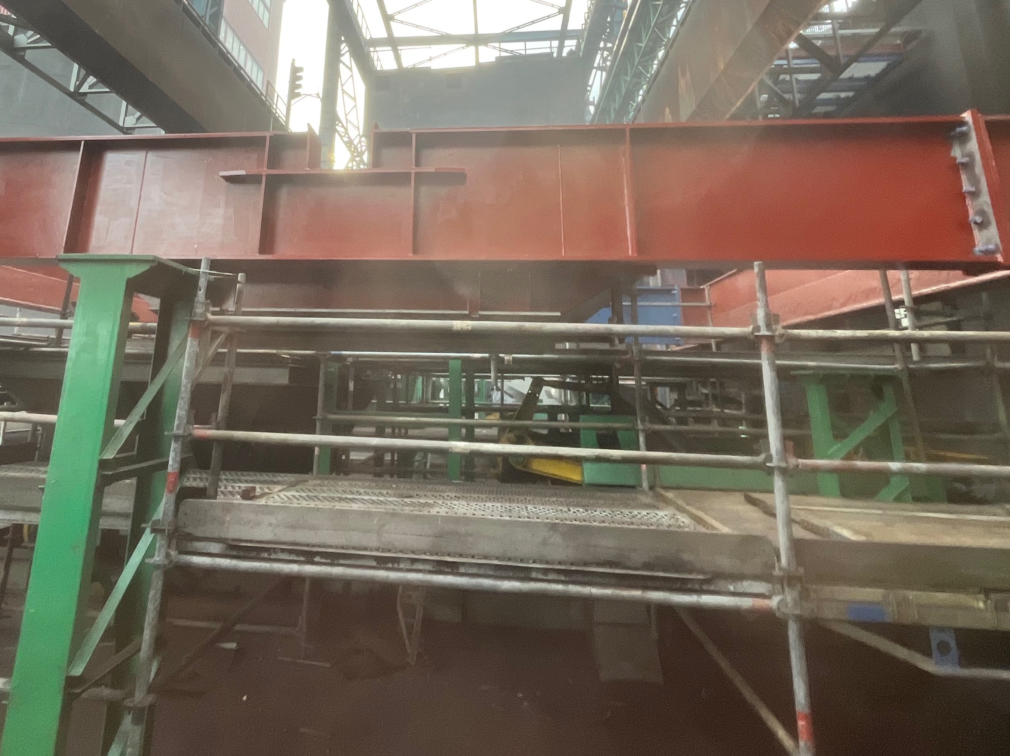 Czerwona, stalowa konstrukcja wsparta na rusztowaniu w hali przemysłowej. Widoczne ślady obróbki i przygotowania do dalszych prac. Ujęcie z dołu.