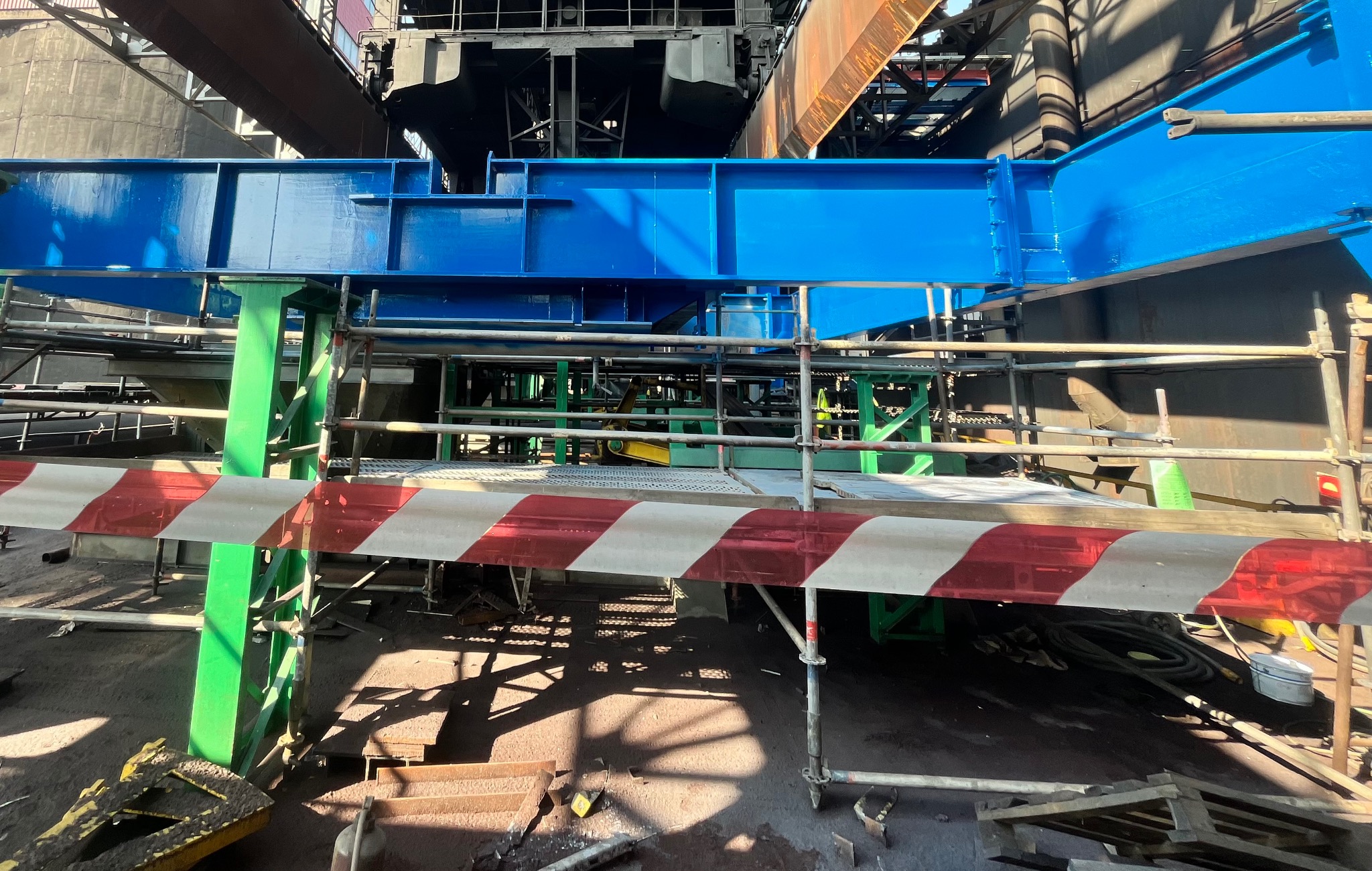 Konstrukcja stalowa w trakcie prac konserwacyjnych, zabezpieczona taśmą ostrzegawczą. Widoczne rusztowania i elementy konstrukcyjne w kolorze niebieskim i zielonym.