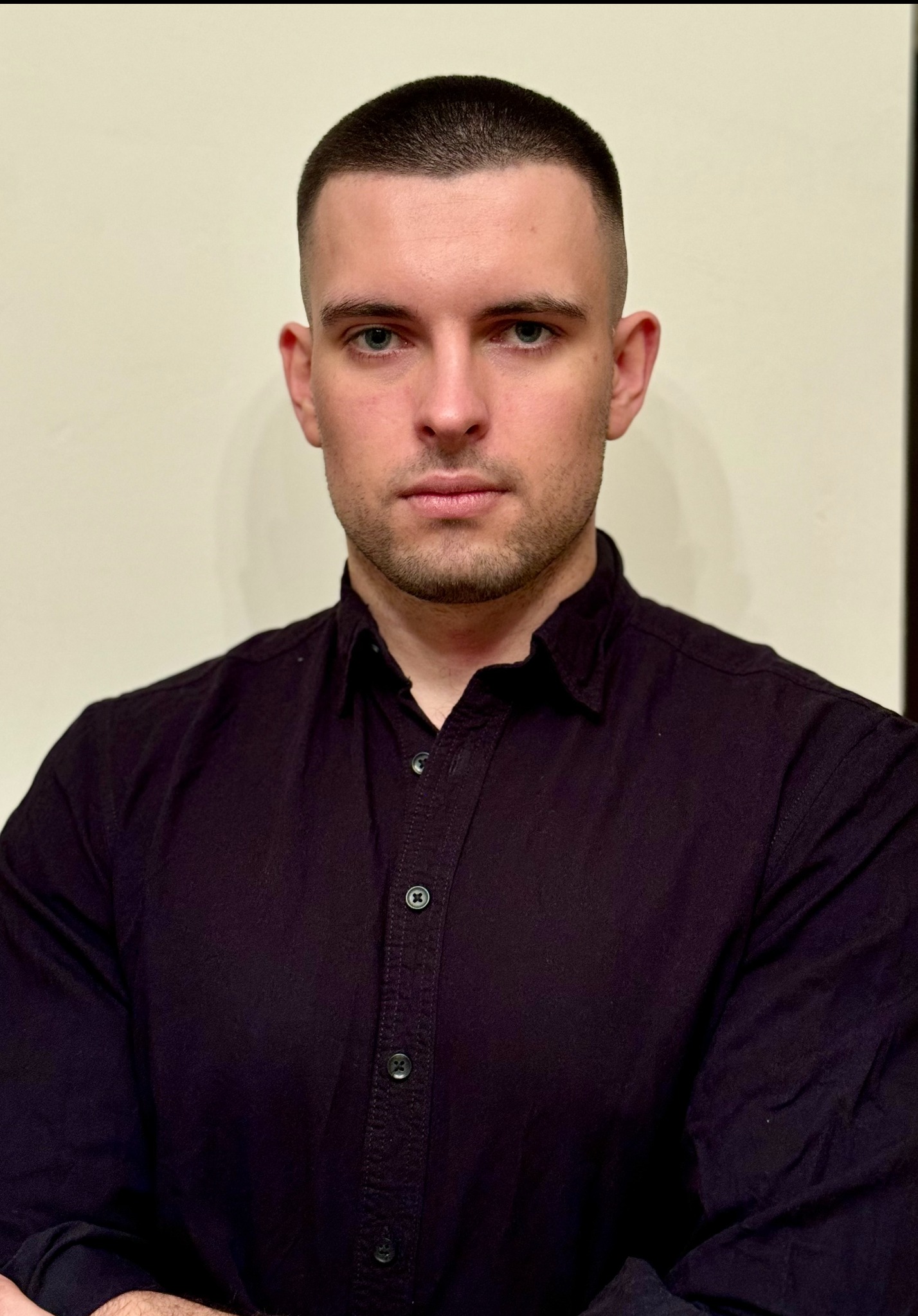 Portret młodego mężczyzny o krótkich włosach, w ciemnej koszuli na tle jasnej ściany. Ujęcie od przodu, z widoczną górną częścią ciała i skrzyżowanymi rękami.