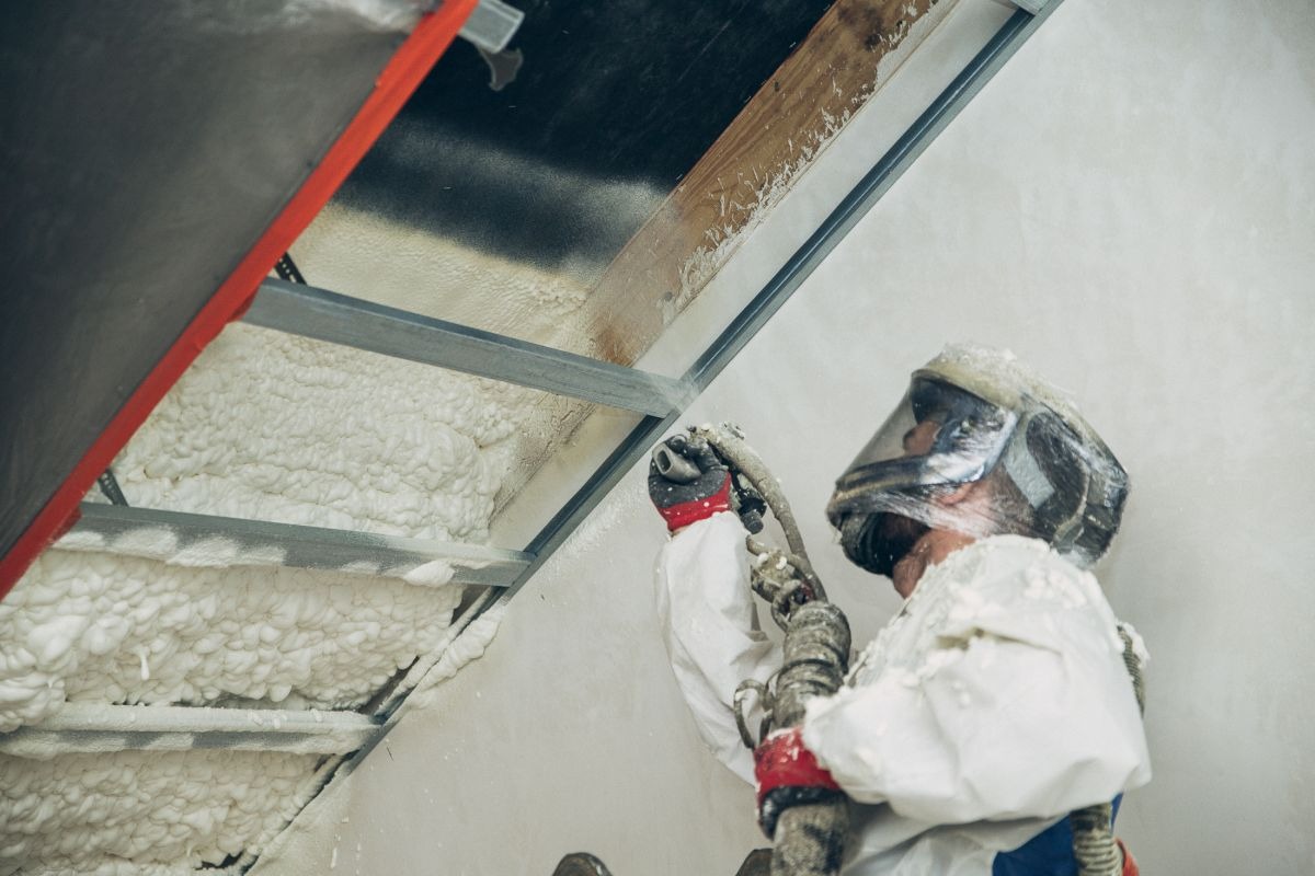 Pracownik w kombinezonie ochronnym aplikuje pianę izolacyjną PUR na metalową konstrukcję poddasza. Widoczne elementy drewniane. Detal ujęcia na pianę i narzędzie.
