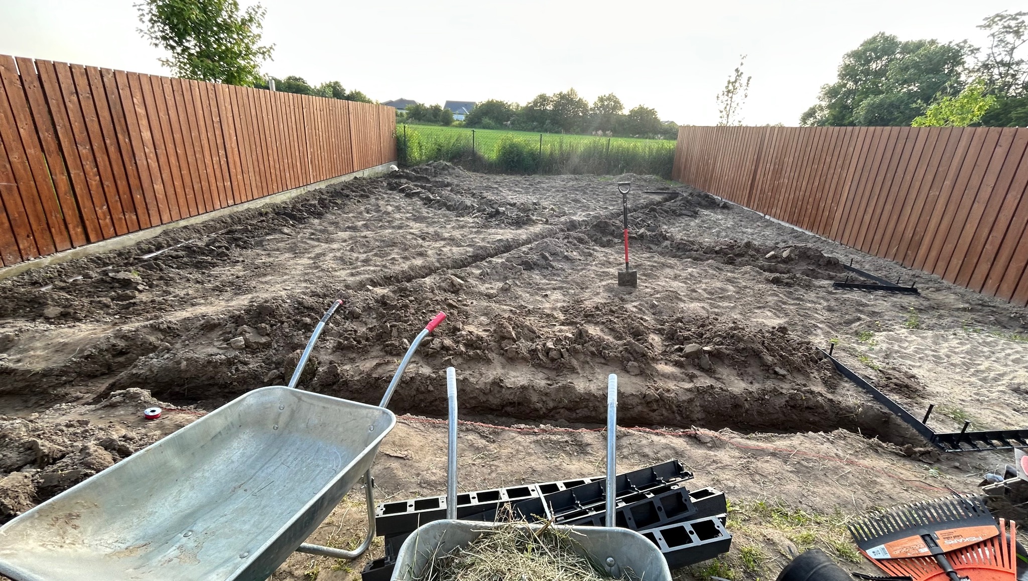 Przygotowany teren pod ogród z wykopanymi rowami pod rabaty, widoczny szpadel, taczka i narzędzia ogrodnicze, ogrodzenie z drewnianych desek w tle.