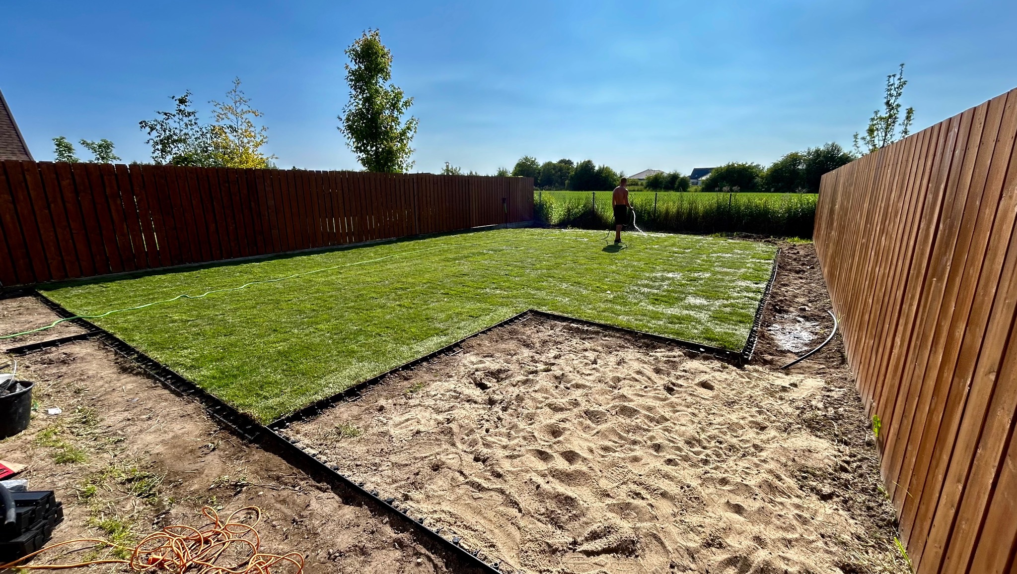Układanie trawy z rolki w ogrodzie. Część trawnika już ułożona, druga część przygotowana podłoże z piasku. Mężczyzna podlewa trawę. W tle drewniany płot.