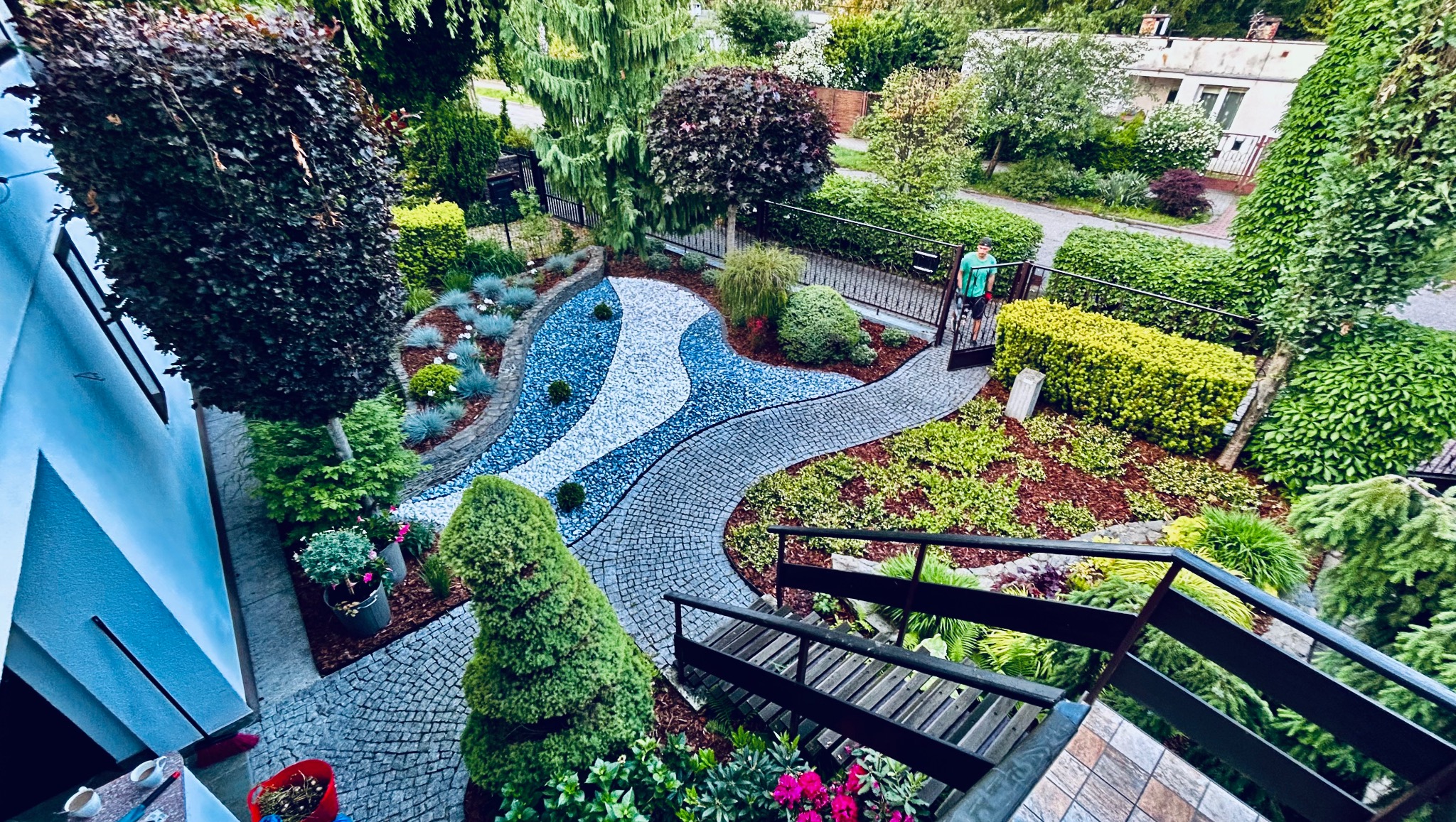 Widok z góry na ogród z ścieżką z kostki, rabatami z kamyków i roślinami ozdobnymi. Osoba w zielonej koszulce wchodzi do ogrodu przez furtkę.