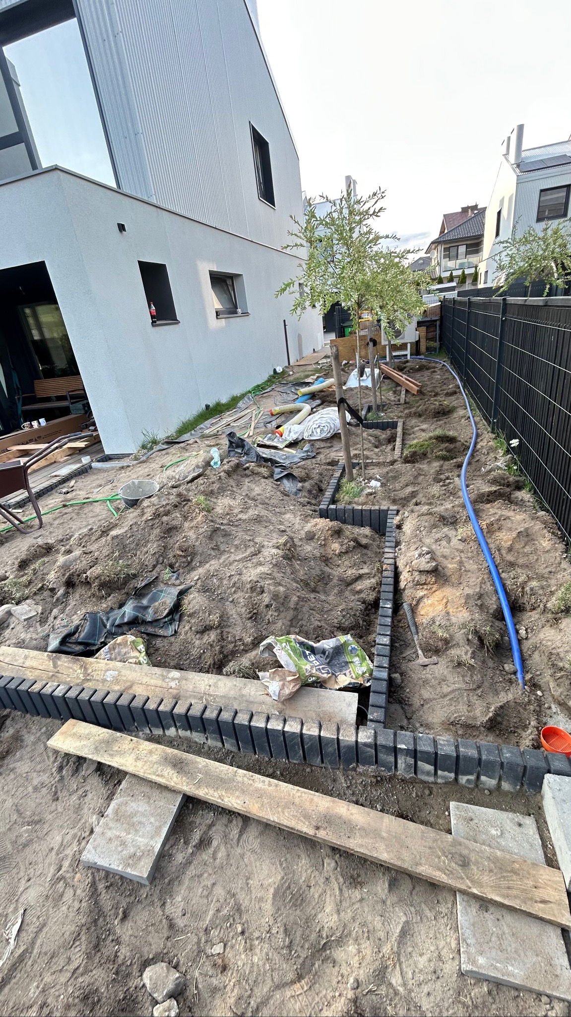 Prace ziemne w ogrodzie: przygotowanie terenu pod nasadzenia, widoczne obrzeża z tworzywa, rozłożona instalacja nawadniająca. W tle nowoczesny dom i ogrodzenie.