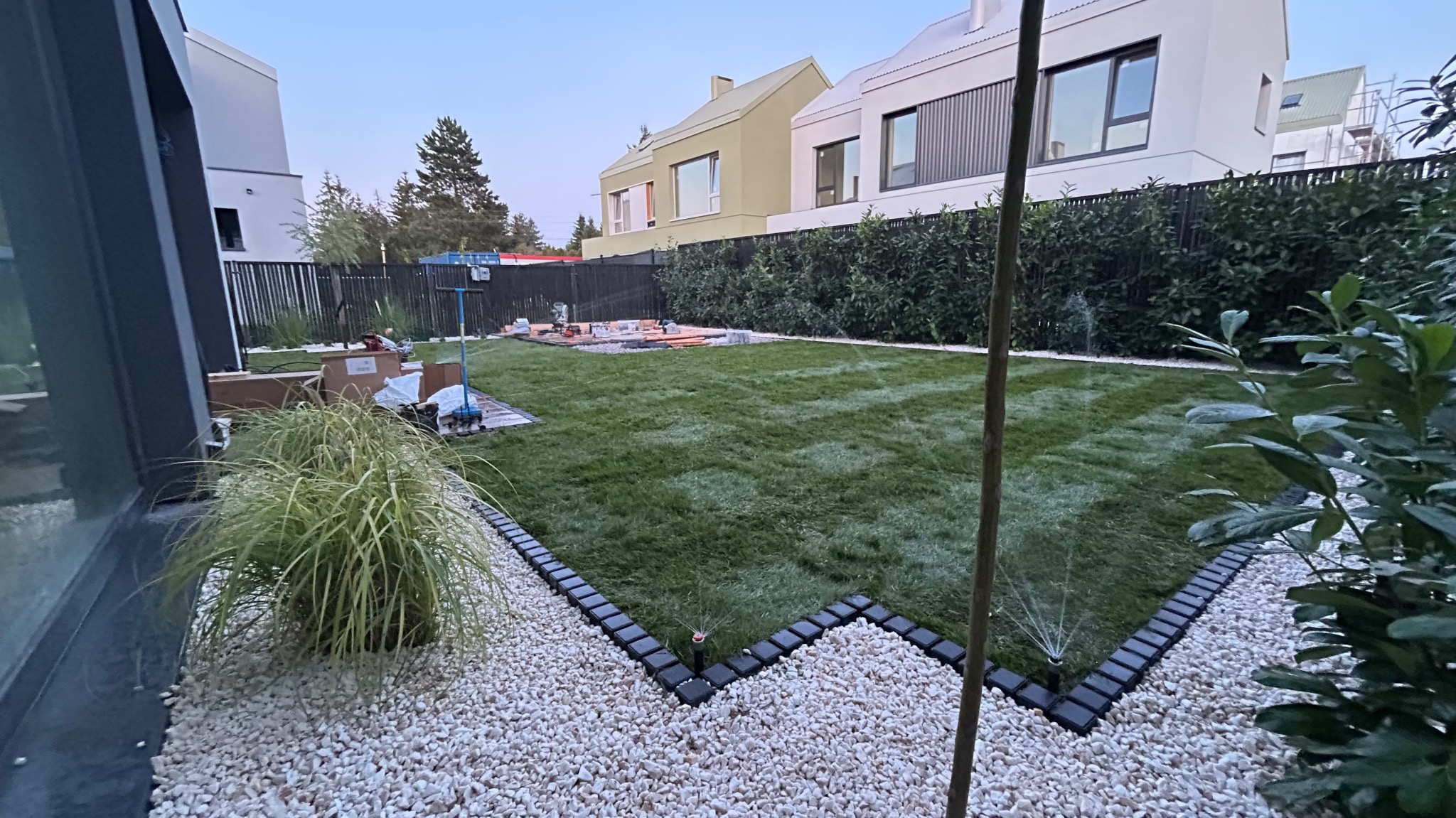 Zadbany trawnik z systemem nawadniania w nowoczesnym ogrodzie. Widoczne zraszacze, obrzeża z kamienia i kostki, w tle domy o minimalistycznej architekturze.