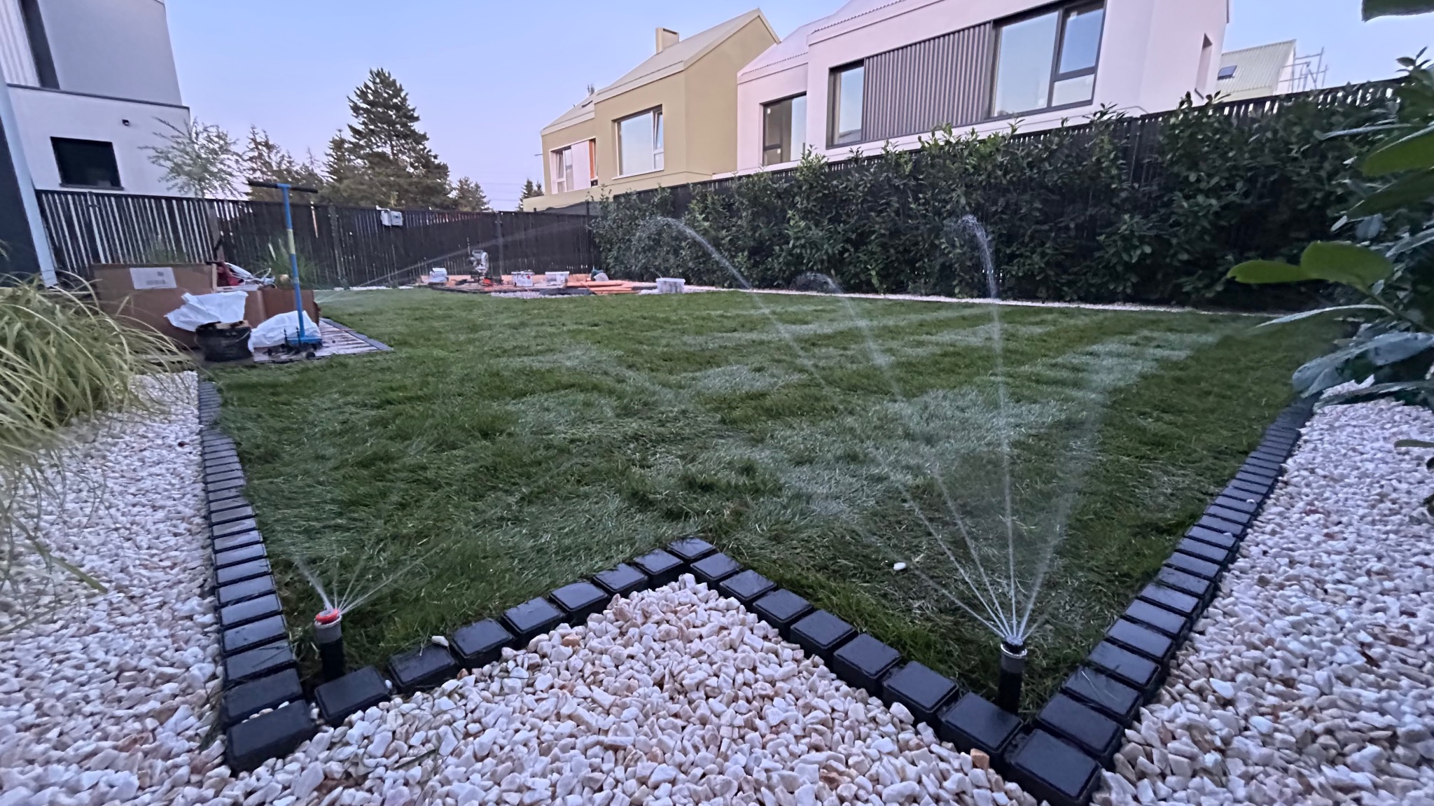 Automatyczny system nawadniania trawnika w nowoczesnym ogrodzie z białymi kamieniami i obrzeżami. Zraszacze w akcji, w tle budynki mieszkalne.