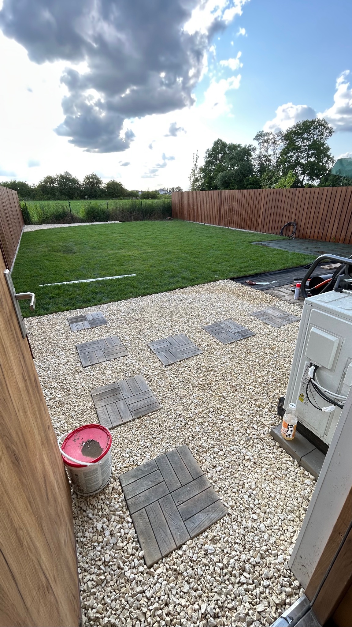 Ogród z trawnikiem, ścieżką z drewnianych płyt na żwirowym podłożu i drewnianym ogrodzeniem. Widoczny kubeł i klimatyzator.