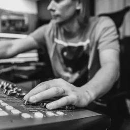 Czarno-białe zbliżenie na rękę mężczyzny z pierścieniem, miksującego dźwięk na profesjonalnym panelu w studiu nagrań. Widoczny profil mężczyzny i fragment klawiatury.