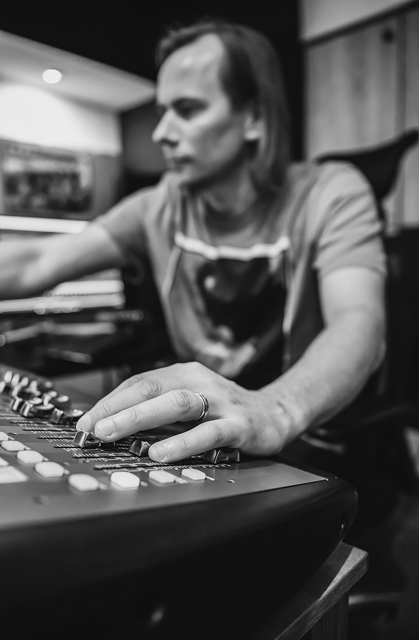 Czarno-białe zbliżenie na rękę mężczyzny z pierścieniem, miksującego dźwięk na profesjonalnym panelu w studiu nagrań. Widoczny profil mężczyzny i fragment klawiatury.