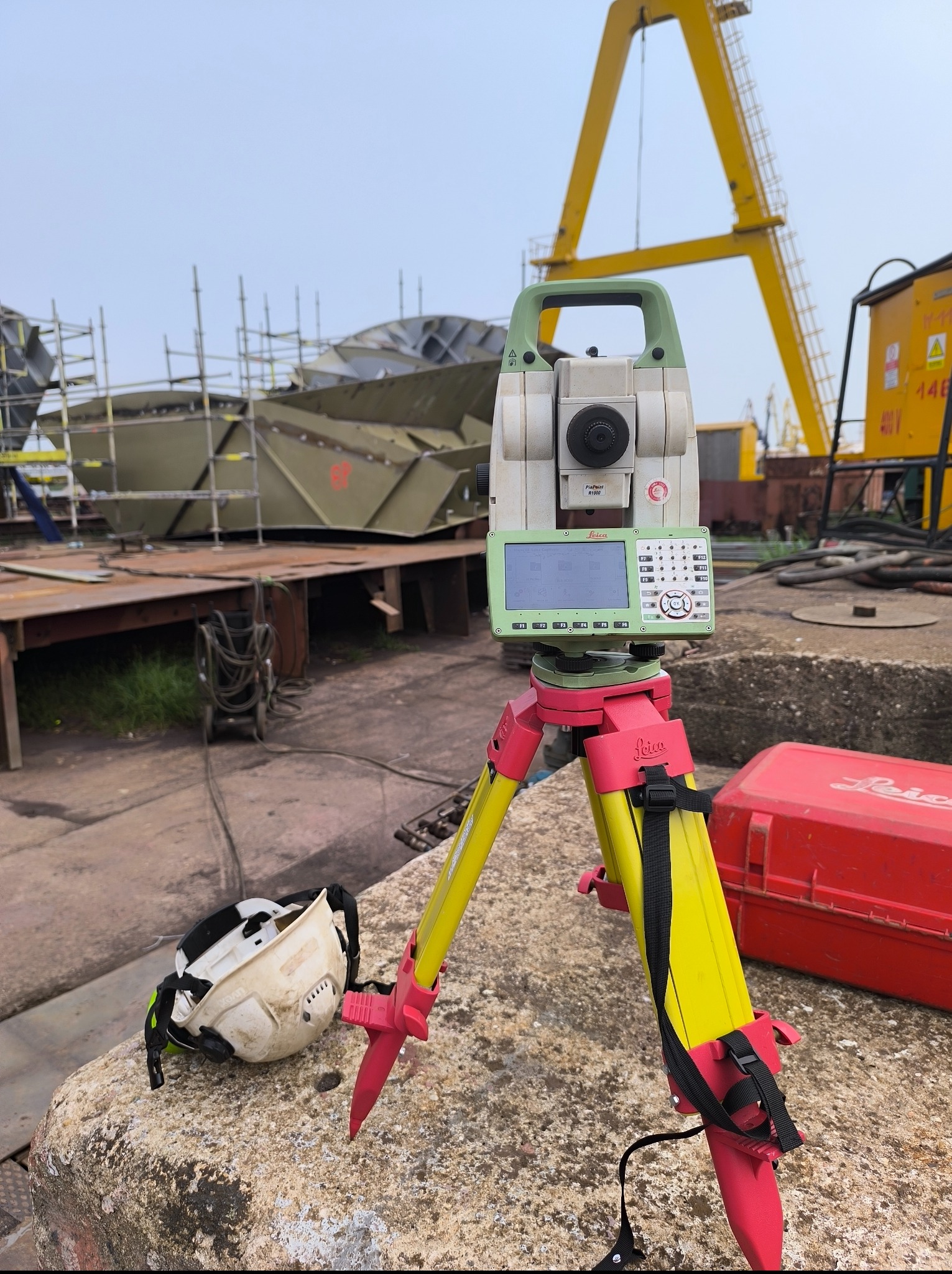 Precyzyjny tachimetr Leica na statywie, na tle konstrukcji stalowej i żurawia, sugerujący pomiary geodezyjne na placu budowy. Obok kask ochronny i czerwona skrzynka narzędziowa.