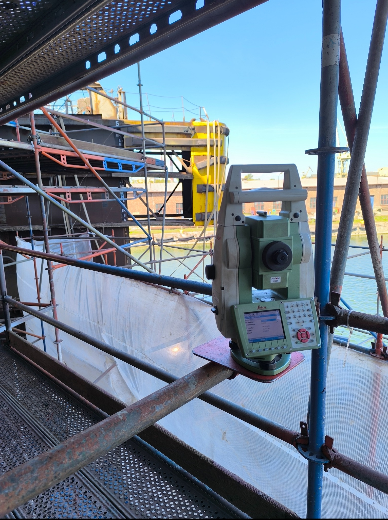 Tacheometr Leica na rusztowaniu, z widokiem na konstrukcję budowlaną i zbiornik wodny w tle, wykonujący pomiary na placu budowy.