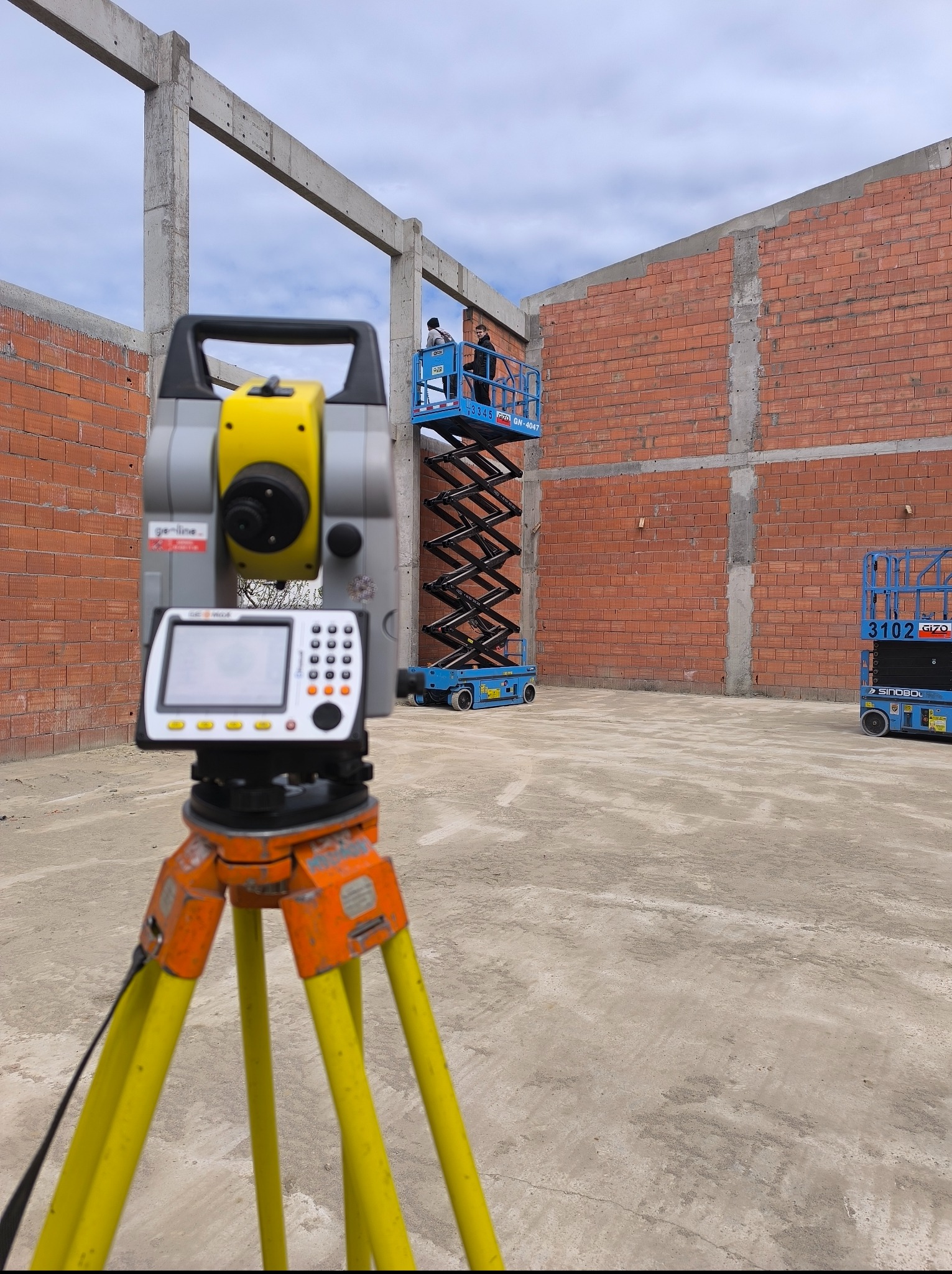 Precyzyjny niwelator na statywie, z żółtymi nogami, na tle budowy z czerwonej cegły i podnośnika koszowego z pracownikami. Pomiar geodezyjny w toku.