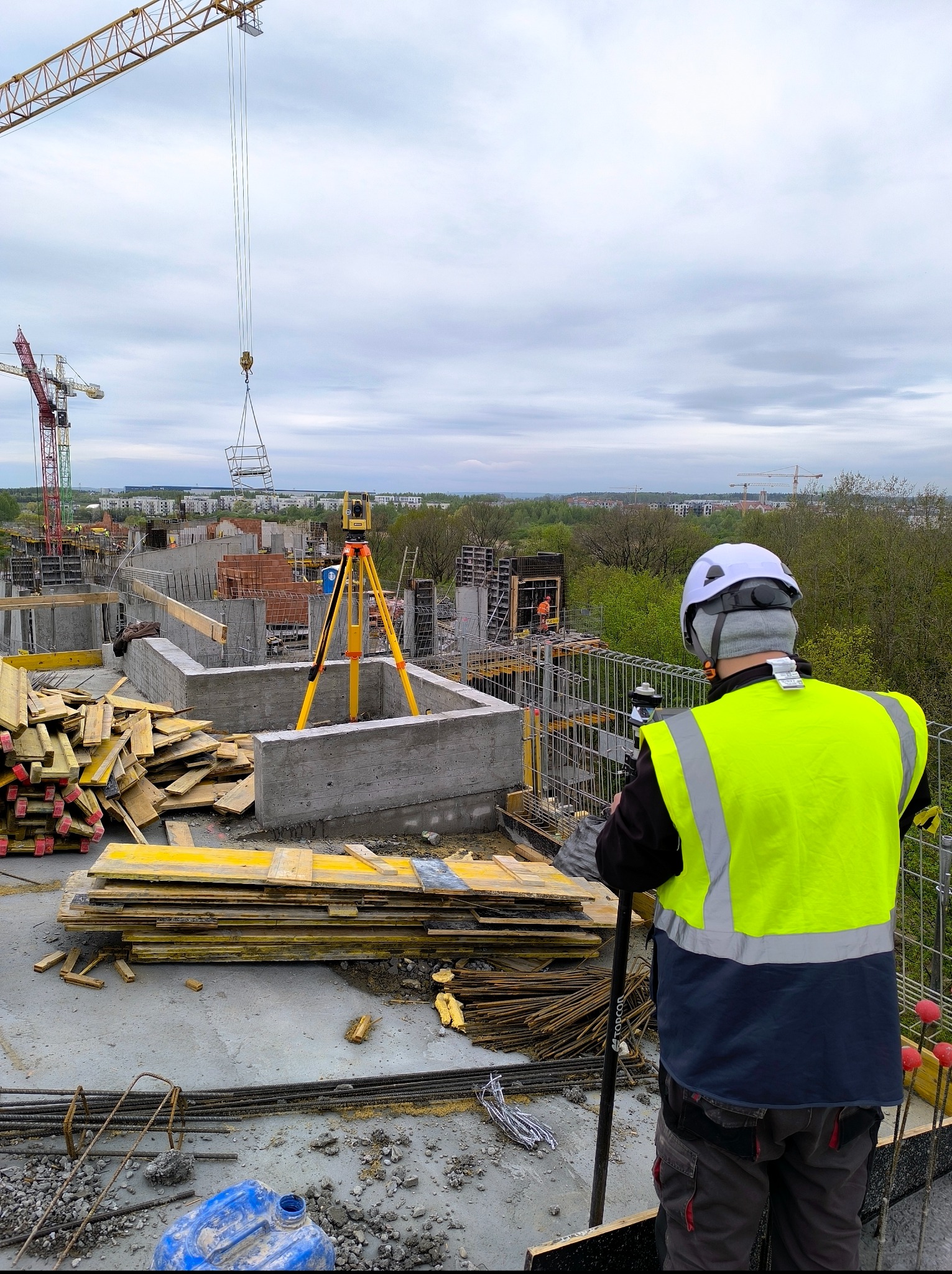 Praca na budowie: osoba w kasku i żółtej kamizelce obsługuje żółty niwelator na tle fundamentów i żurawi, z widocznym stosem drewnianych desek.