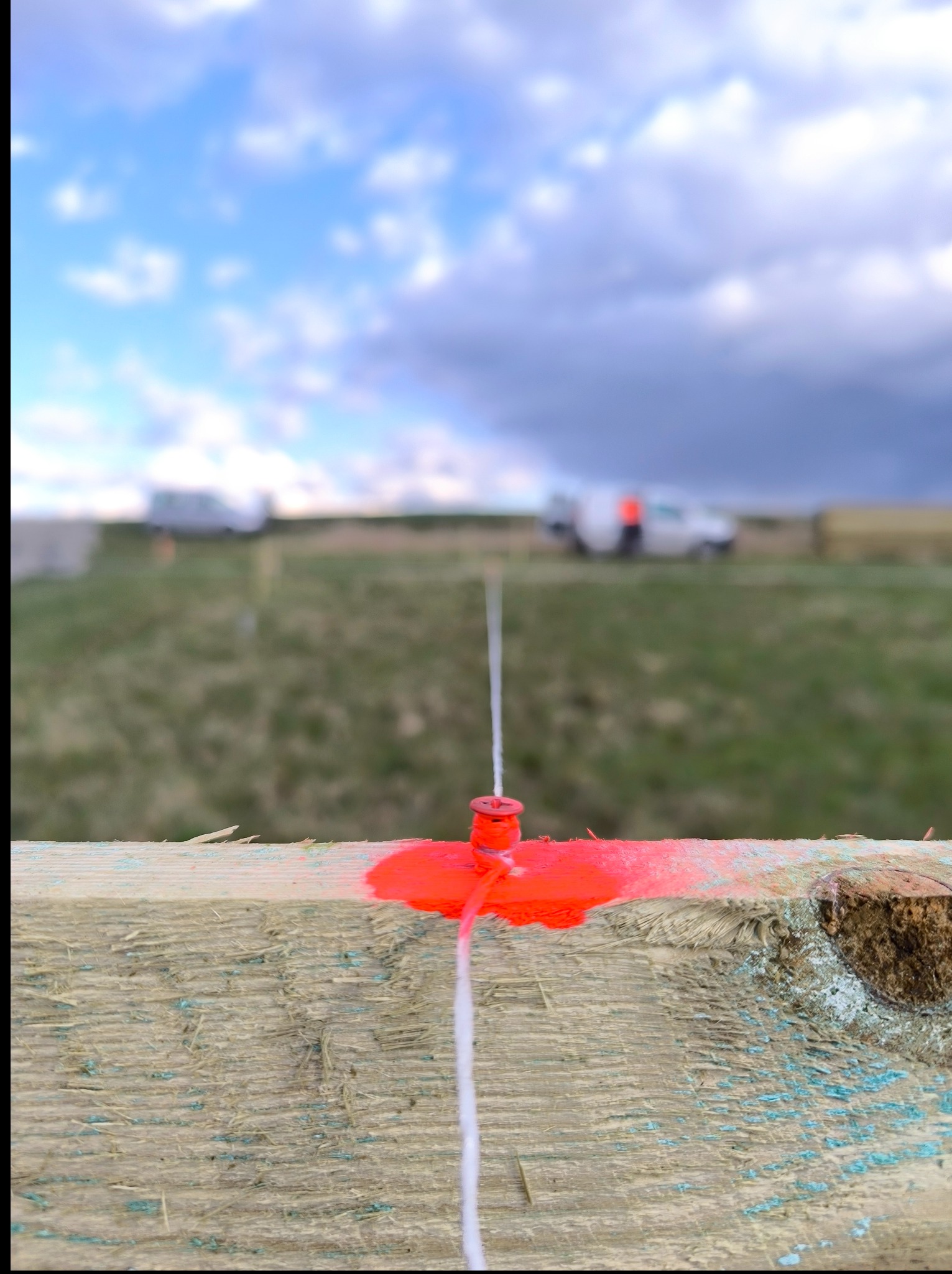Czerwony kołek z linką na drewnianej desce, w tle łąka i niebo. Wyznaczanie terenu.
