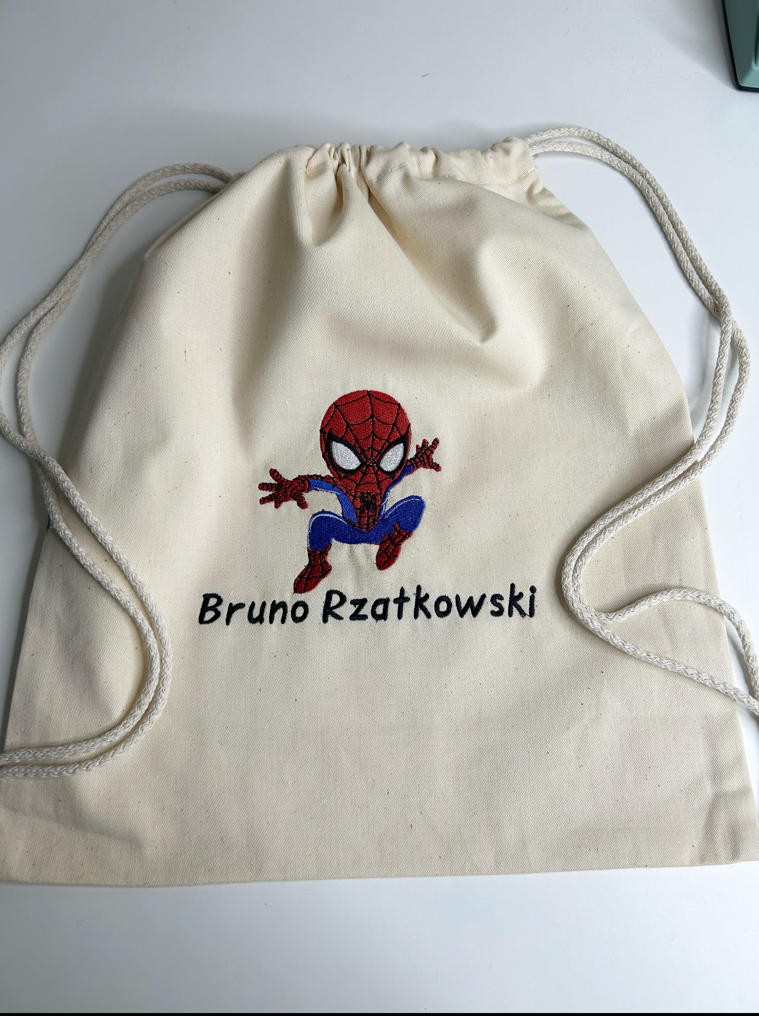 Beżowy worek z haftowanym Spider-Manem i imieniem 'Bruno Rzatkowski', leżący na białym tle. Widoczne sznurki ściągające worek.