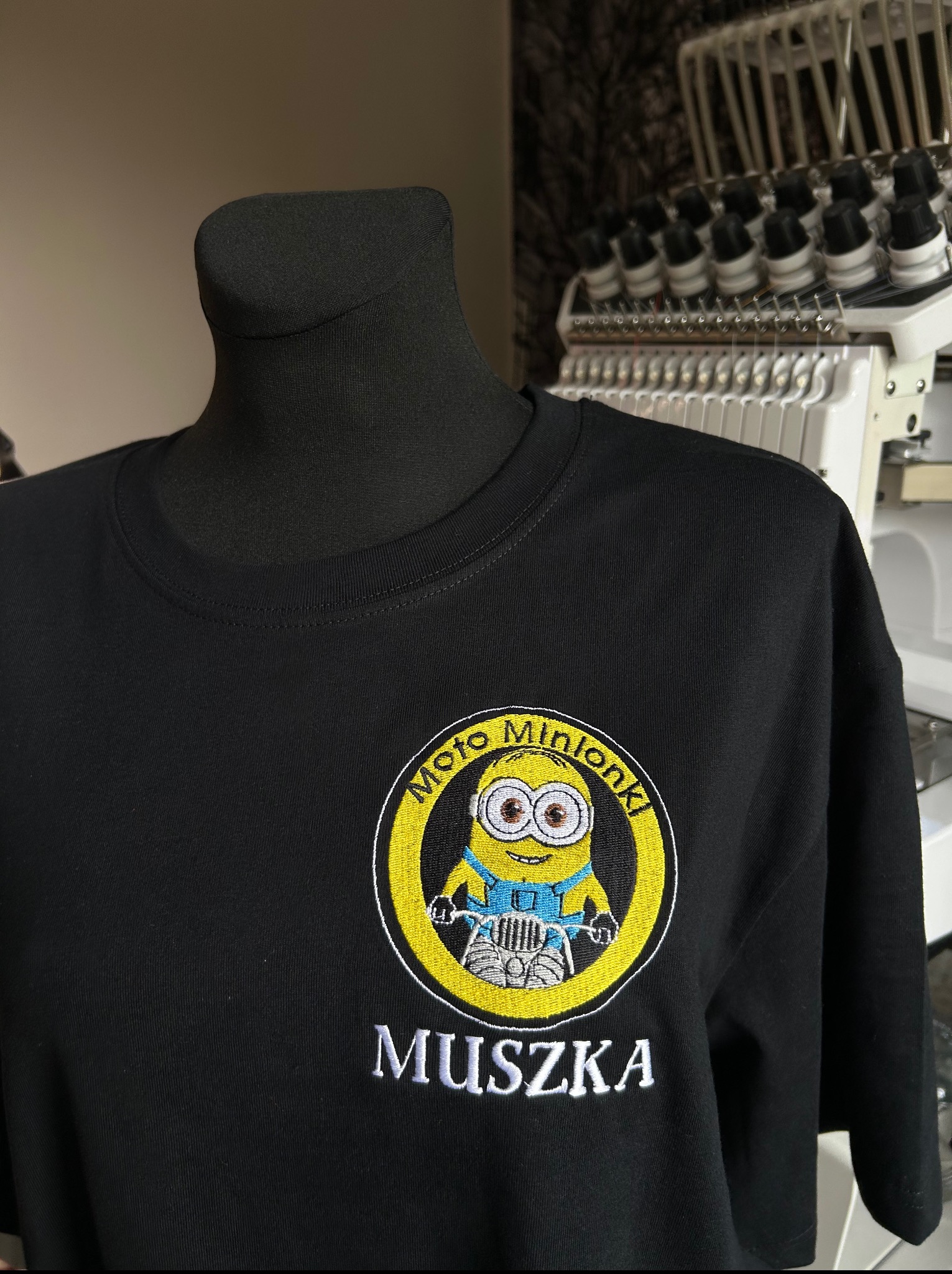 Czarna koszulka z haftowanym logo 'Moto Minionki' i napisem 'Muszka', prezentowana na manekinie krawieckim. W tle widoczna maszyna do haftowania.