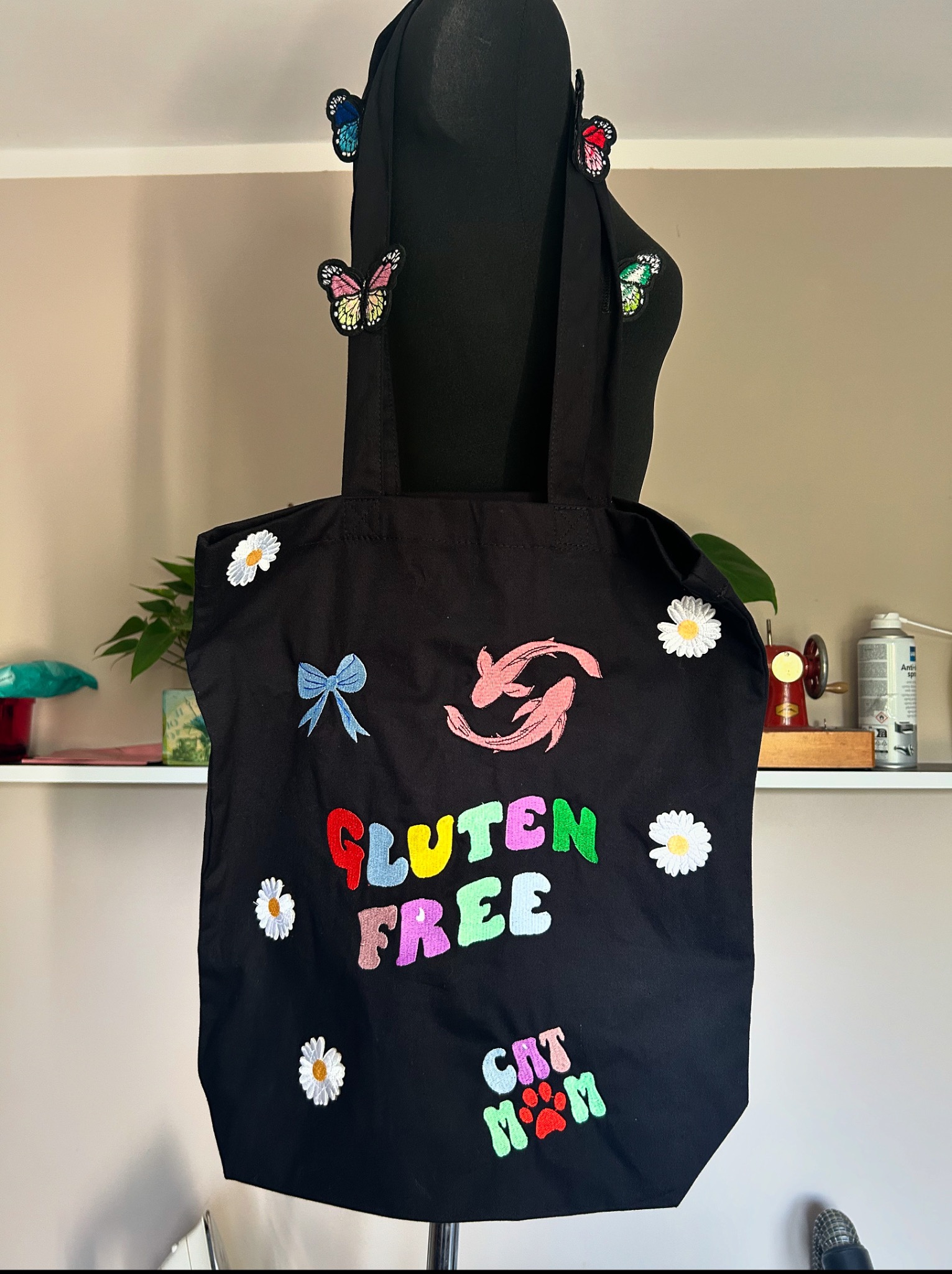 Czarna torba bawełniana z kolorowymi haftami (kwiaty, napisy, rybki koi) w stylu DIY, wisząca na manekinie z motylami, na tle półki z roślinami i maszyną do szycia.