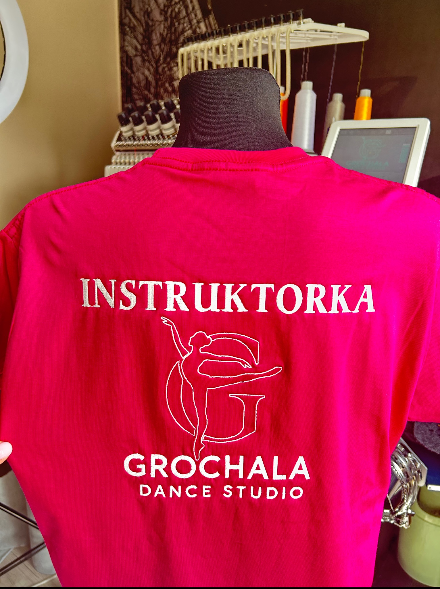 Różowa koszulka z białym haftem: 'INSTRUKTORKA', logo taneczne i 'GROCHALA DANCE STUDIO'. Na manekinie, obok maszyna do haftowania.
