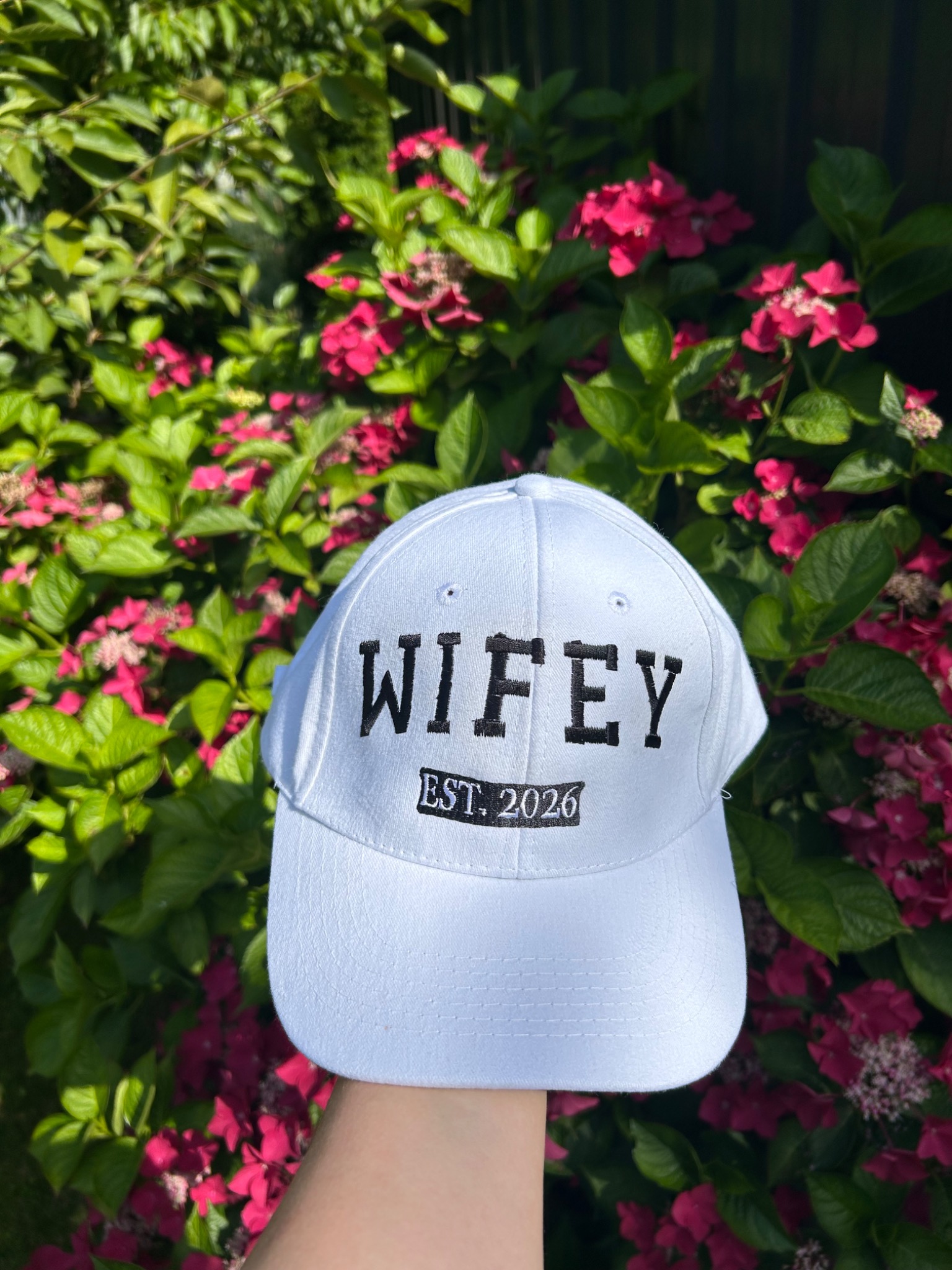 Biała czapka z daszkiem, haftowany napis 'WIFEY EST. 2026', trzymana na tle kwitnącego krzewu. Słoneczny dzień, delikatne cieniowanie.