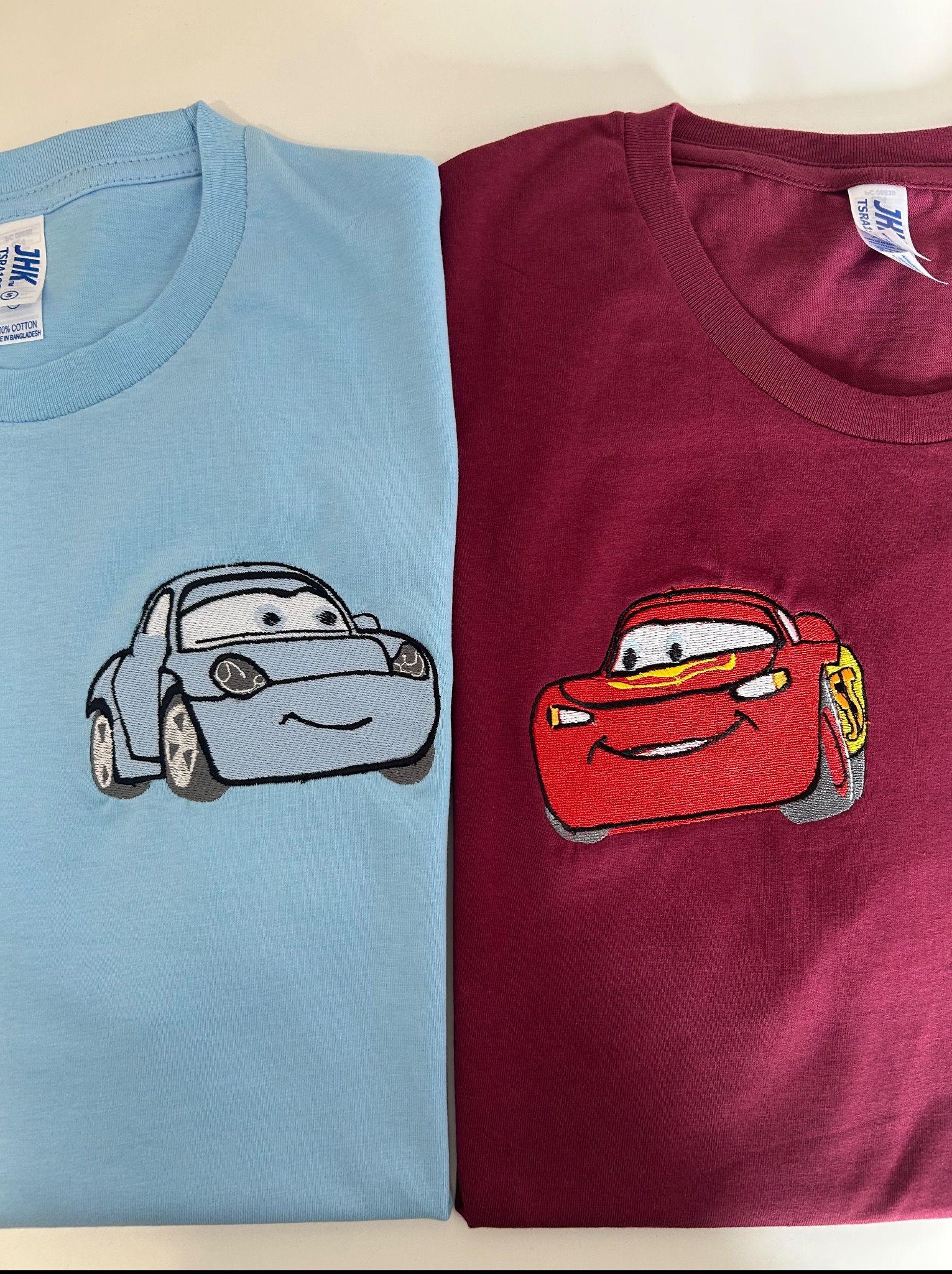 Dwa t-shirty: błękitny z haftem Sally i bordowy z haftem Zygzaka McQueen, postacie z filmu Auta, leżą obok siebie na białym tle.