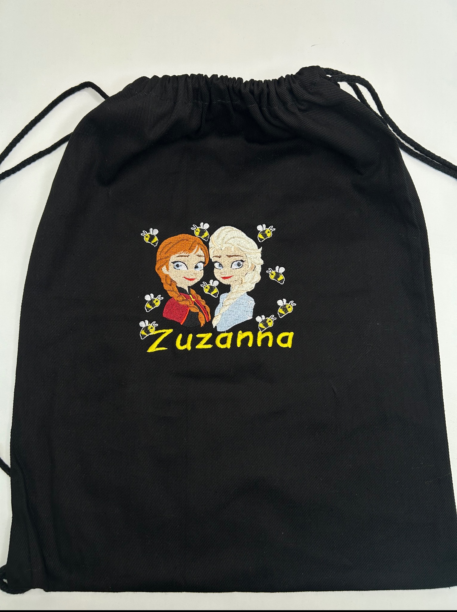 Czarny worek ze sznurkiem, ozdobiony haftem postaci z bajki i imieniem 'Zuzanna', otoczonych motywem pszczół. Detal haftu wykonany z dbałością o szczegóły.