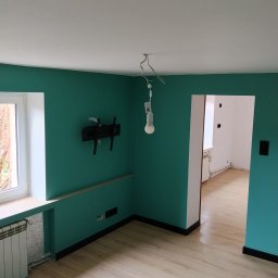 Dominik Rozanski - Pokój po remoncie: turkusowe ściany, jasna podłoga, białe okno i sufit. Widoczny grzejnik, uchwyt na TV i wisząca żarówka. W tle widok na kolejne, białe pomieszczenie.