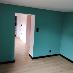Dominik Rozanski - Pomalowane na turkusowo ściany w pokoju z jasną podłogą i czarnymi listwami przypodłogowymi. Widoczne gniazdka elektryczne i przejście do innego pomieszczenia.