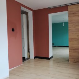 Dominik Rozanski - Wnętrze po malowaniu ścian na ceglasty kolor i położeniu jasnej podłogi. Widoczne drzwi przesuwne i fragment ściany w odcieniu turkusowym w kolejnym pomieszczeniu.