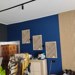 Dominik Rozanski - Niebieska ściana z trzema dekoracyjnymi panelami z geometrycznym wzorem, drewniana ściana z pionowymi listwami i fragment pokoju z widocznym grzejnikiem.