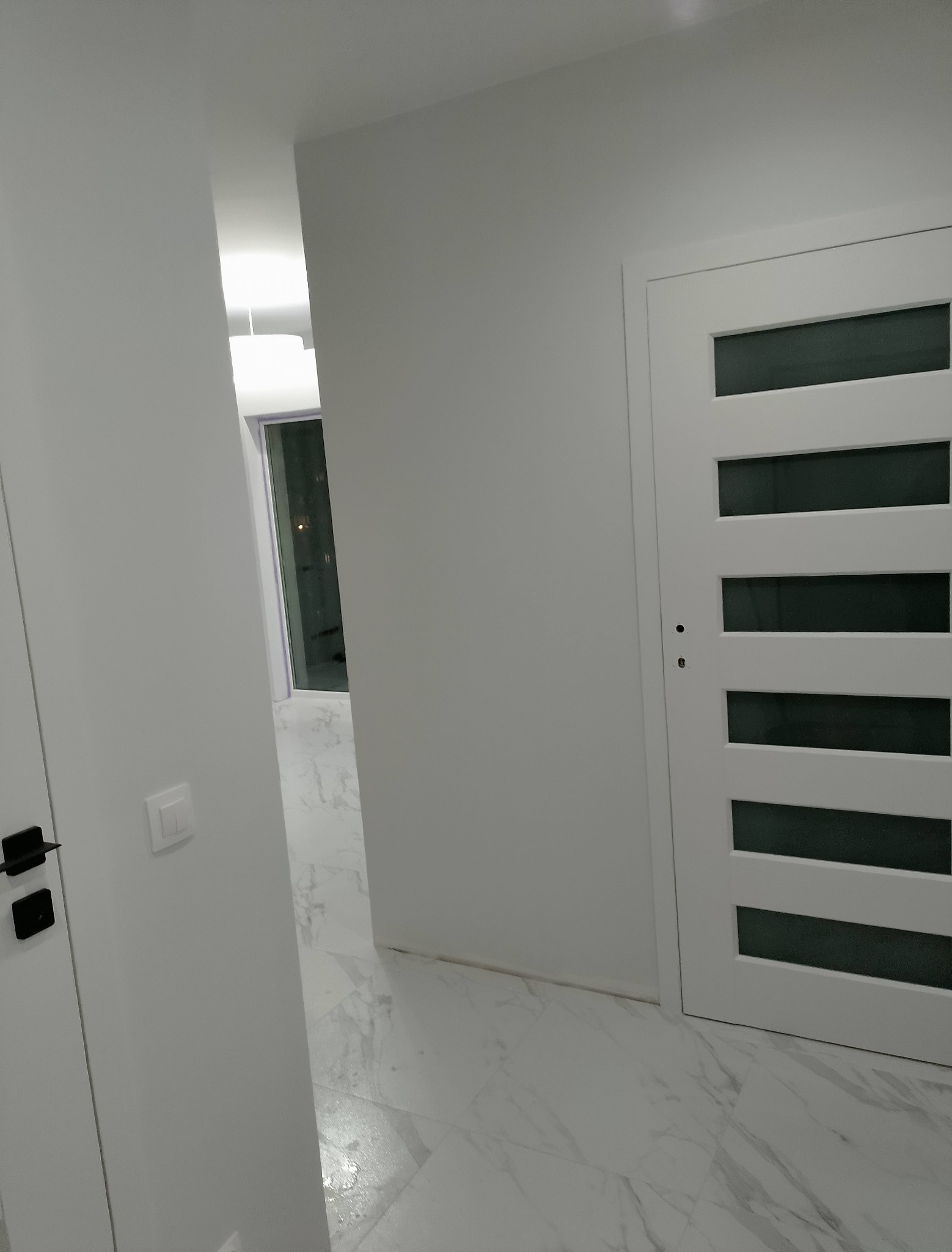 Białe wnętrze: korytarz z marmurową podłogą, białe drzwi z przeszkleniami i widok na oświetlony pokój. Minimalistyczny design, nowoczesne wykończenie.