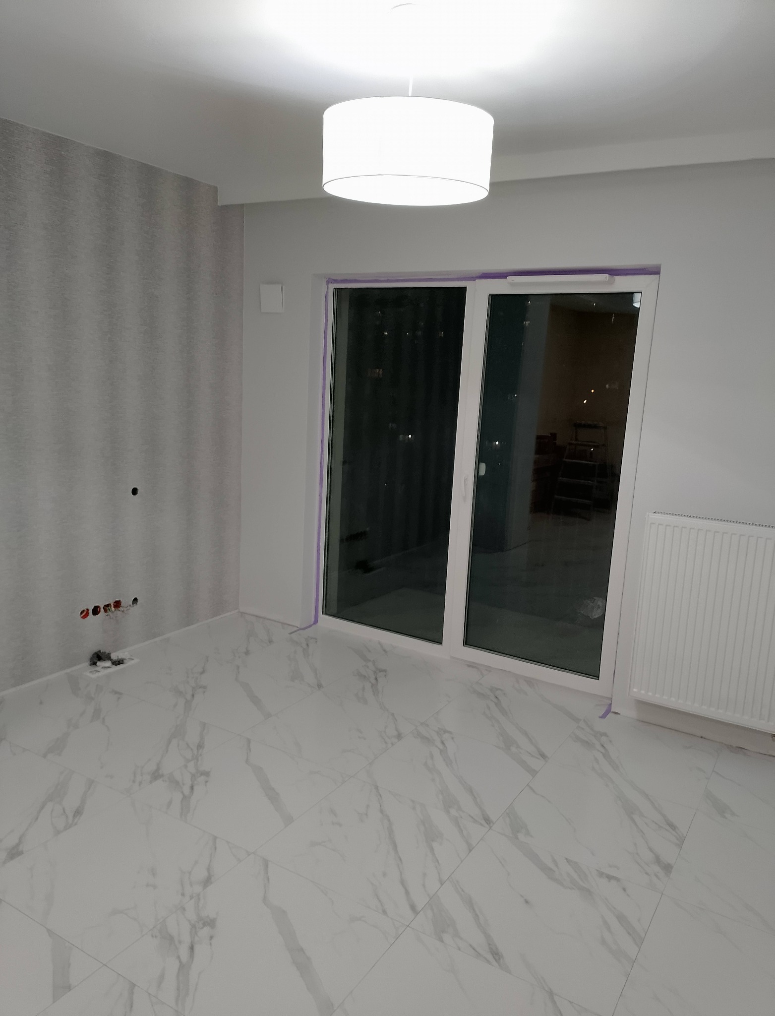 Jasne wnętrze z marmurową podłogą i przesuwnymi drzwiami balkonowymi. Widoczne przygotowane gniazdka elektryczne i grzejnik. Minimalistyczna lampa sufitowa.
