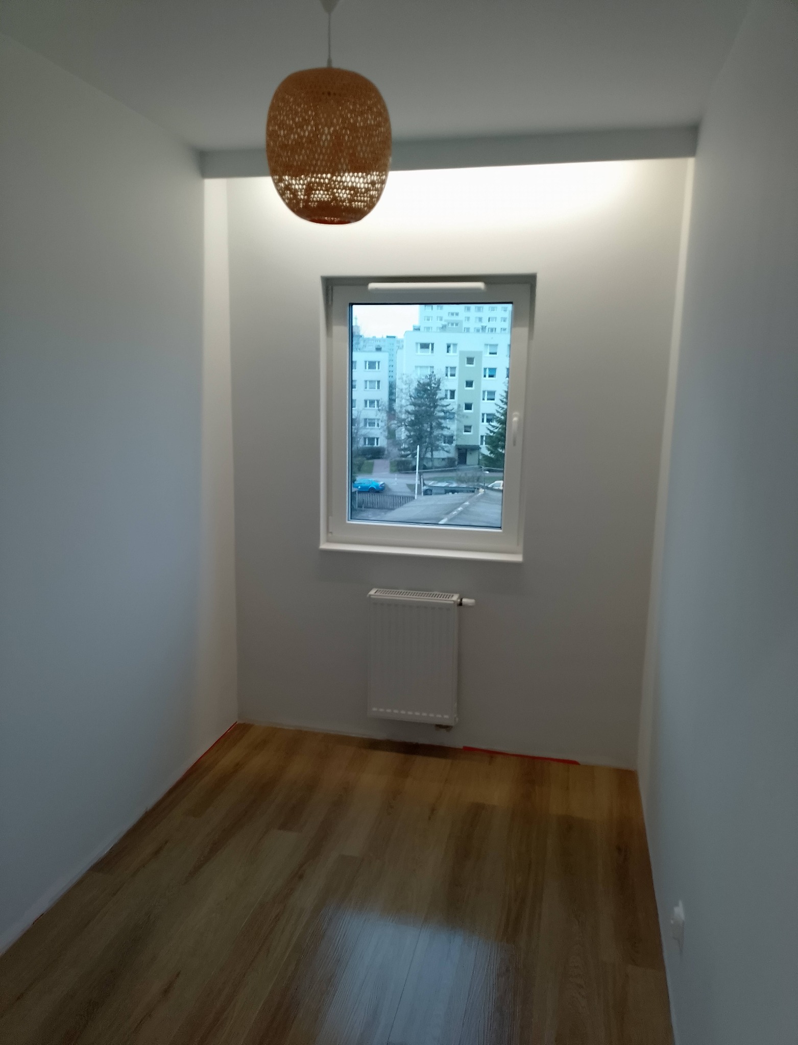 Jasne wnętrze po remoncie: drewniana podłoga, białe ściany, okno z widokiem na blok i pleciona lampa. Minimalistyczny design, świeżo po wykończeniu.