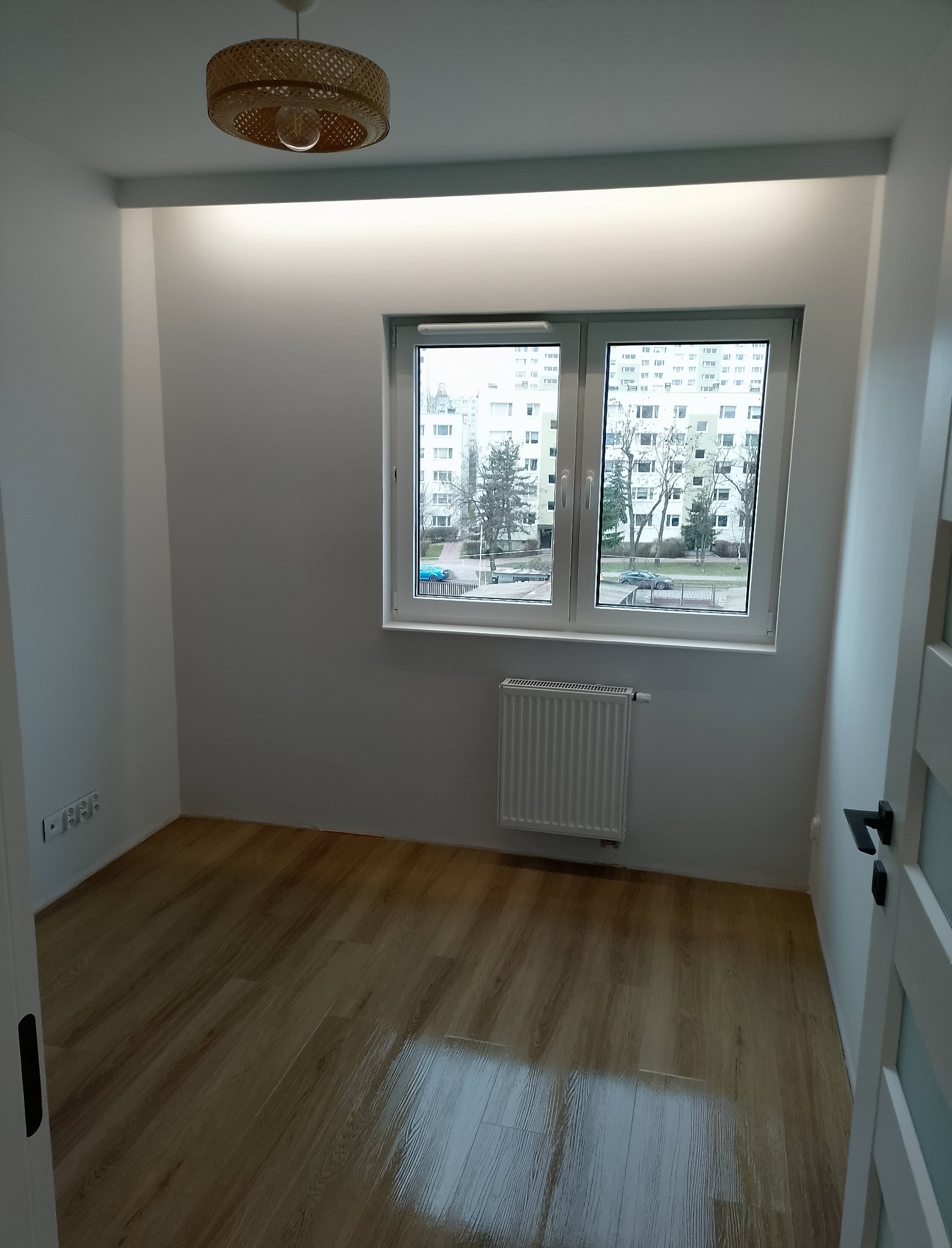 Jasne wnętrze pokoju po remoncie: drewniana podłoga, białe ściany, okno z widokiem na blok, grzejnik i lampa z wikliny. Minimalistyczny design.