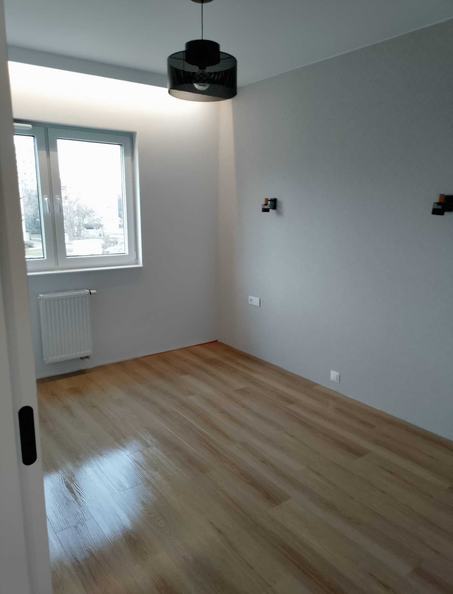 Pusty pokój po remoncie: parkiet, szare ściany z gniazdkami, okno z grzejnikiem, czarna lampa. Widok zza otwartych drzwi, podkreślający świeżość wykończenia.