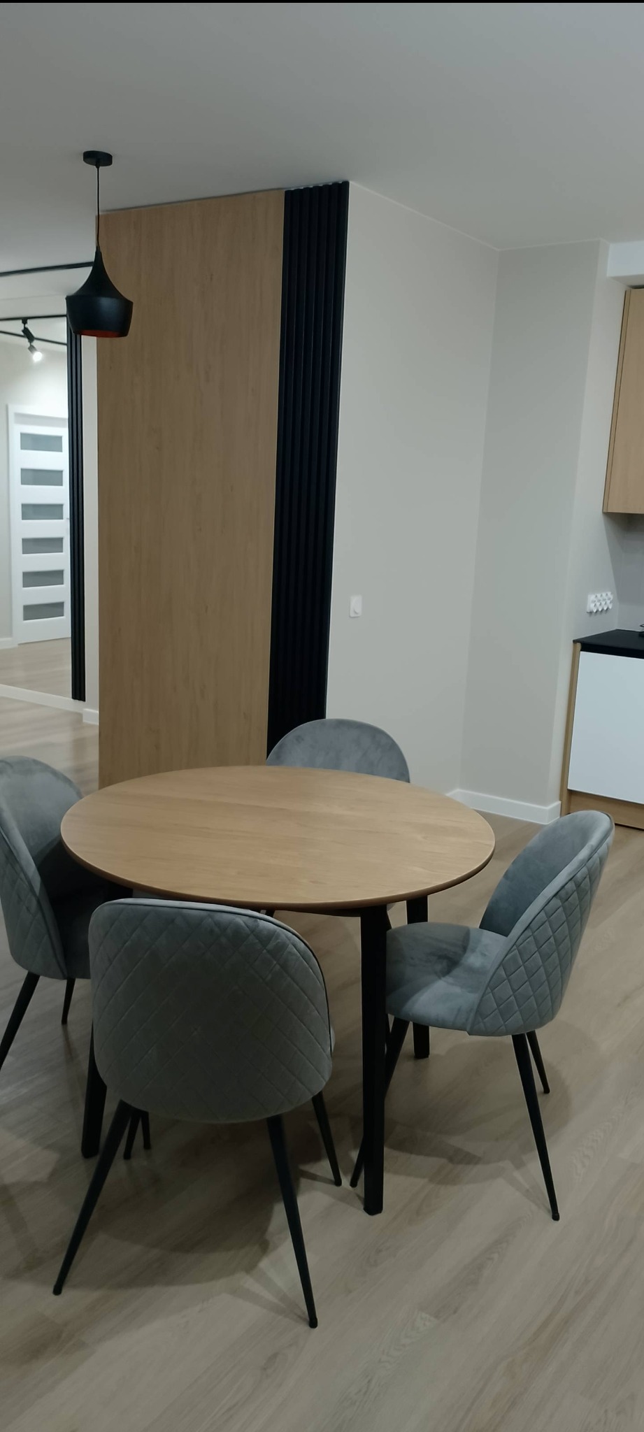 Nowoczesna jadalnia z okrągłym, drewnianym stołem i czterema szarymi krzesłami z pikowanymi oparciami. W tle drewniana ścianka działowa z czarnymi lamelami.
