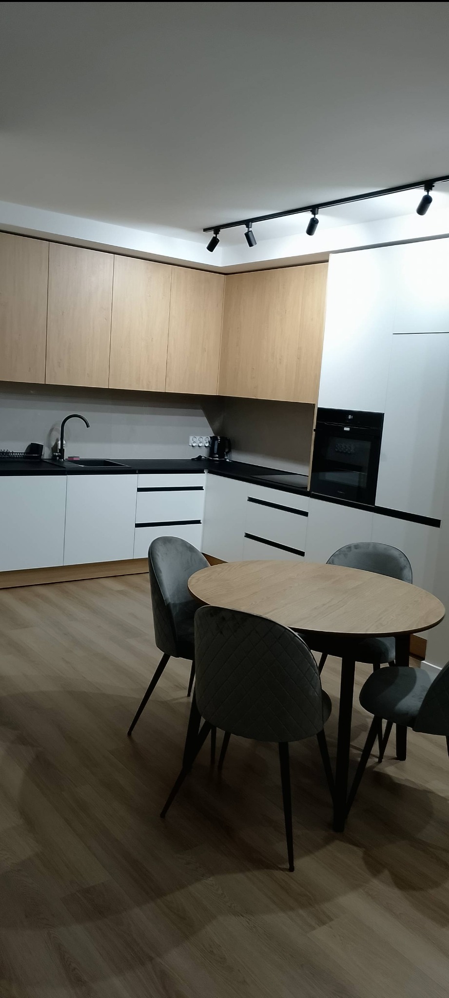 Nowoczesna kuchnia z drewnianymi i białymi szafkami, czarnym blatem i okrągłym stołem z szarymi krzesłami. Oświetlenie punktowe na suficie.