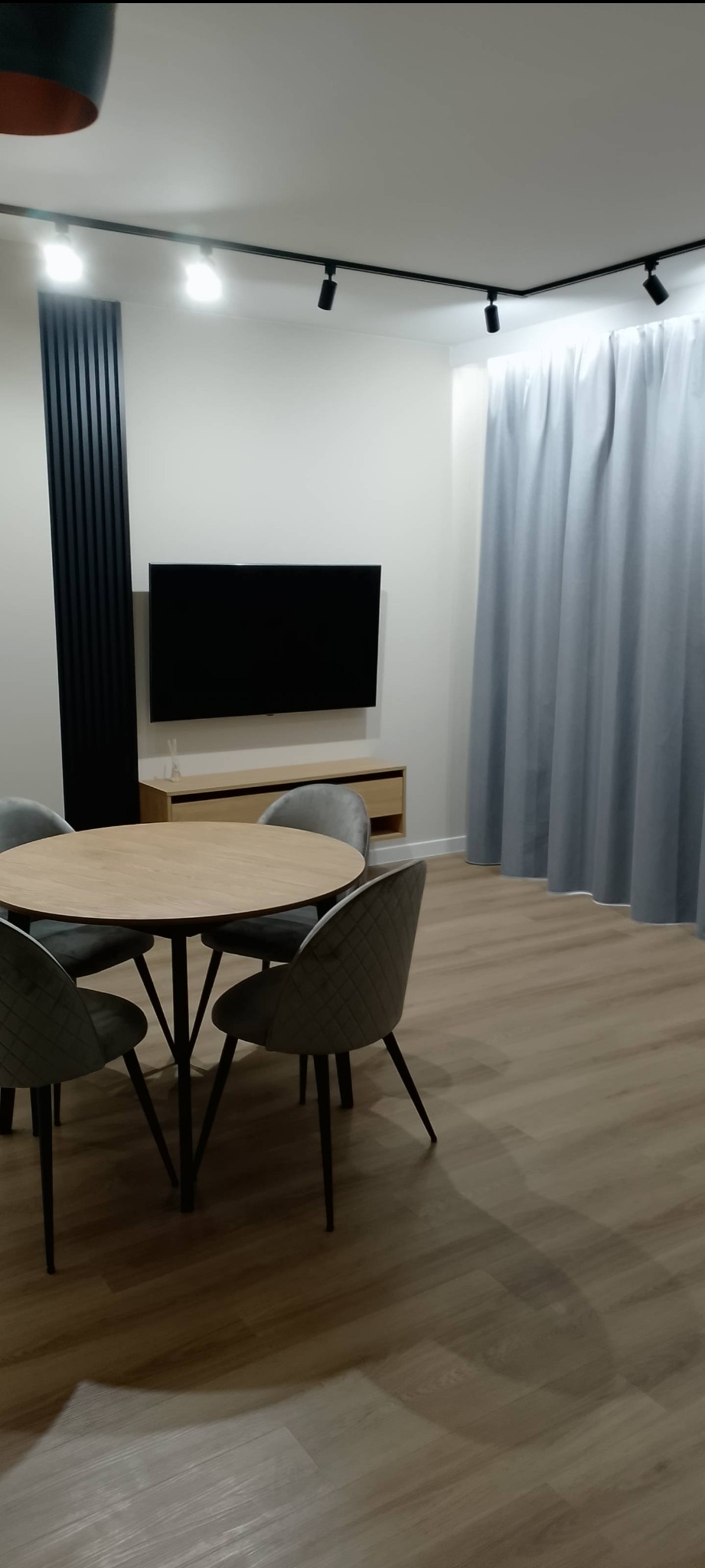 Nowoczesny salon z okrągłym stołem, szarymi krzesłami i minimalistycznym oświetleniem szynowym. Na ścianie telewizor i dekoracyjny panel z lameli.
