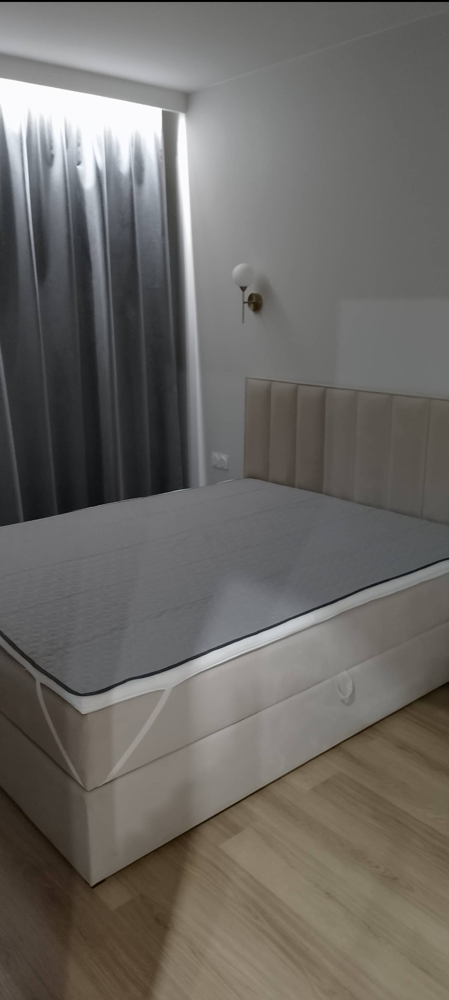 Beżowe łóżko kontynentalne z szarym materacem i wezgłowiem w minimalistycznej sypialni z zasłonami i lampą ścienną.