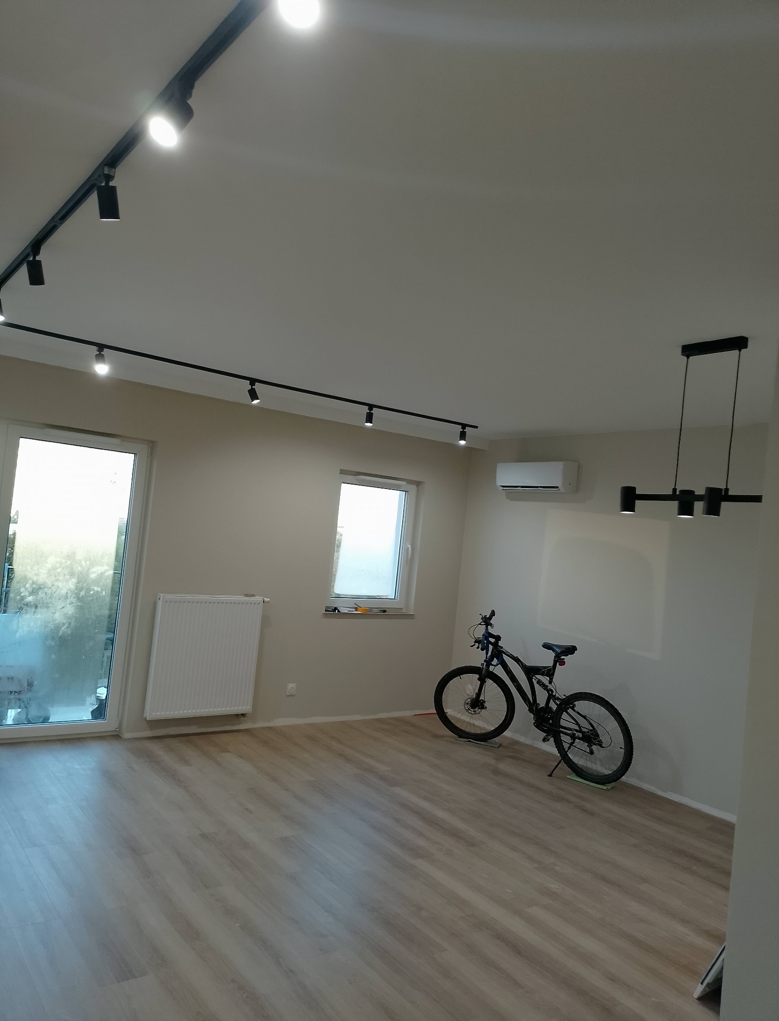 Jasne wnętrze po remoncie w Łodzi: panele podłogowe, biały grzejnik, okno, klimatyzacja i czarne oświetlenie szynowe. Minimalistyczny design.