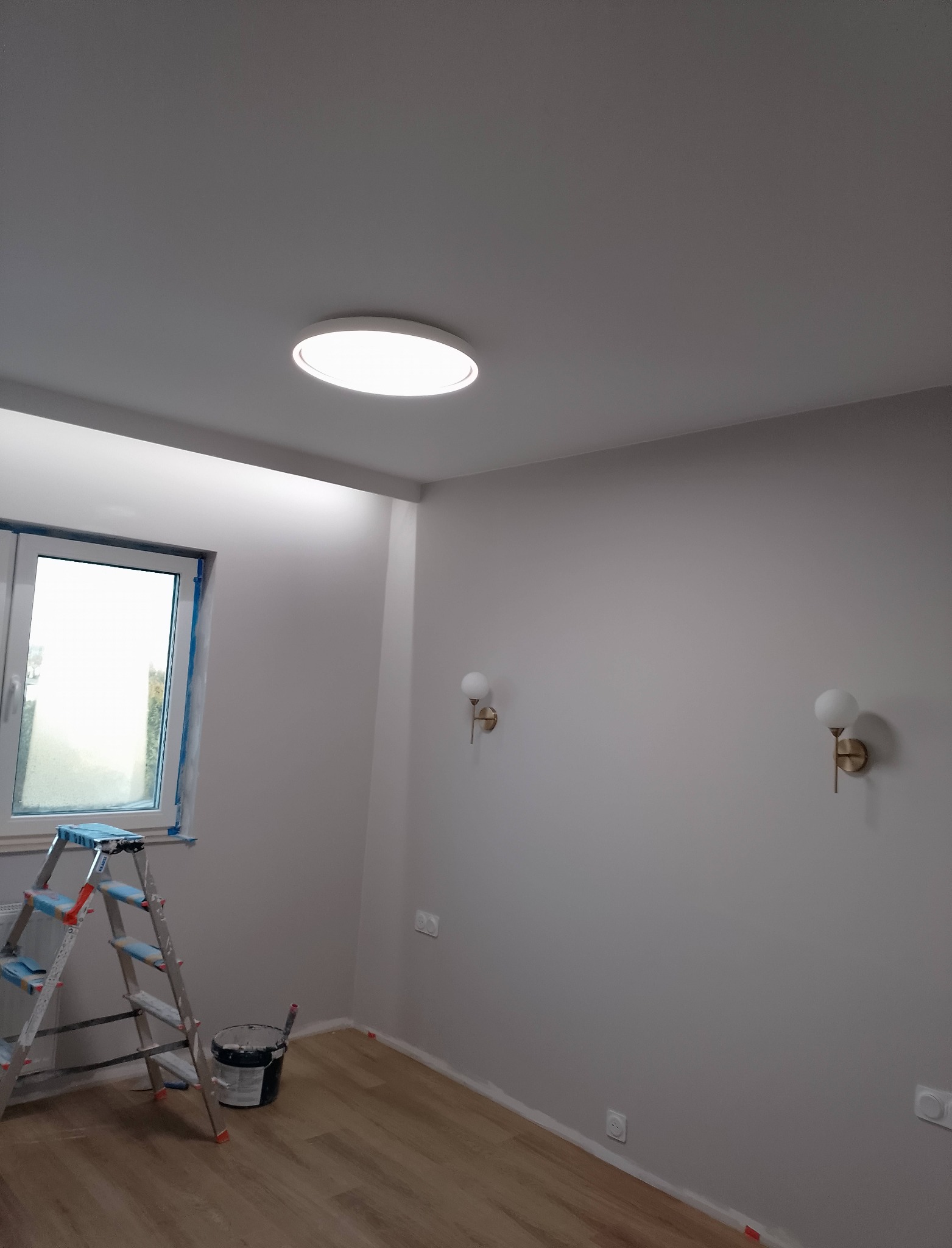 Pokój po malowaniu ścian na szaro, widoczne lampy ścienne, okno z zabezpieczeniem, drabina i wiadro z farbą. Podłoga drewniana.